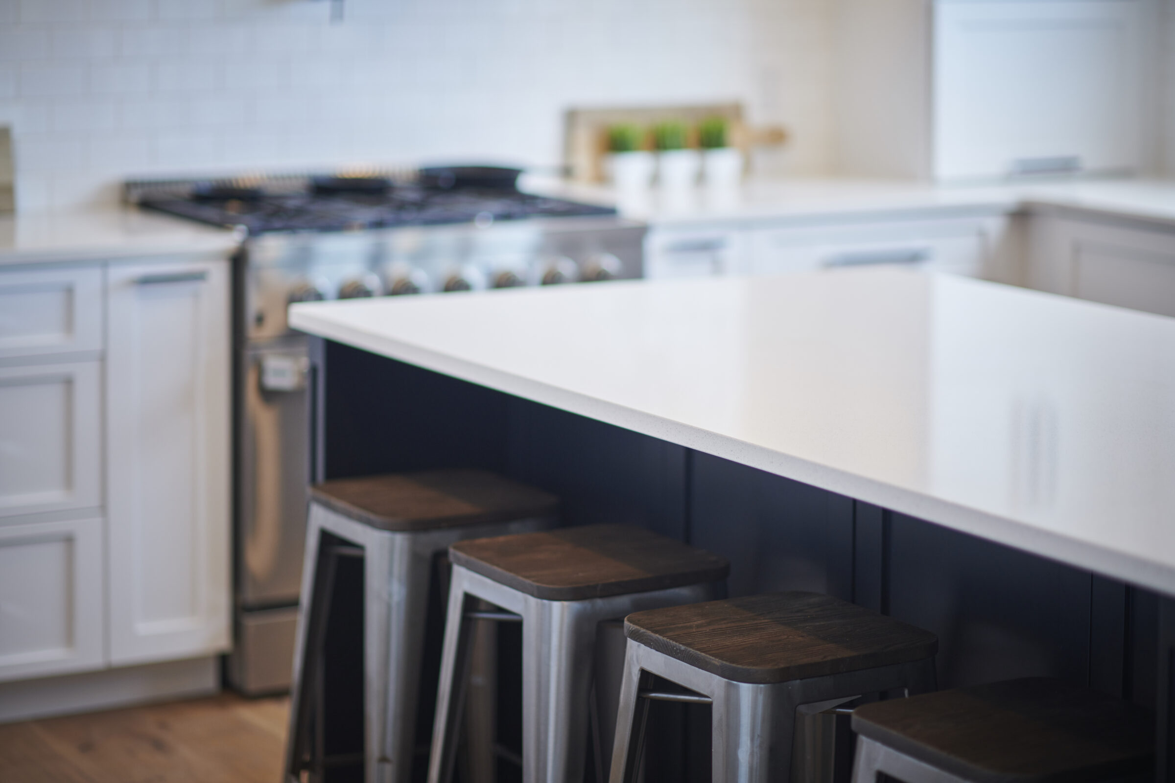 Modern kitchen interior with white countertops, dark cabinetry, metallic stools, and a gas stove. Clean, stylish design with bright lighting and a tiled backsplash.