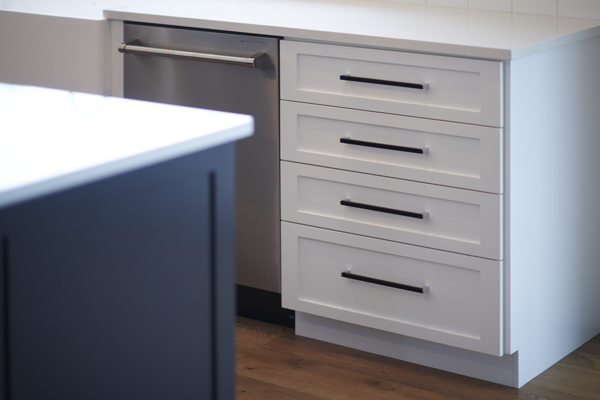 Modern kitchen cabinet with four white drawers, featuring sleek black handles, against a white countertop and gray-blue cabinetry to the left.