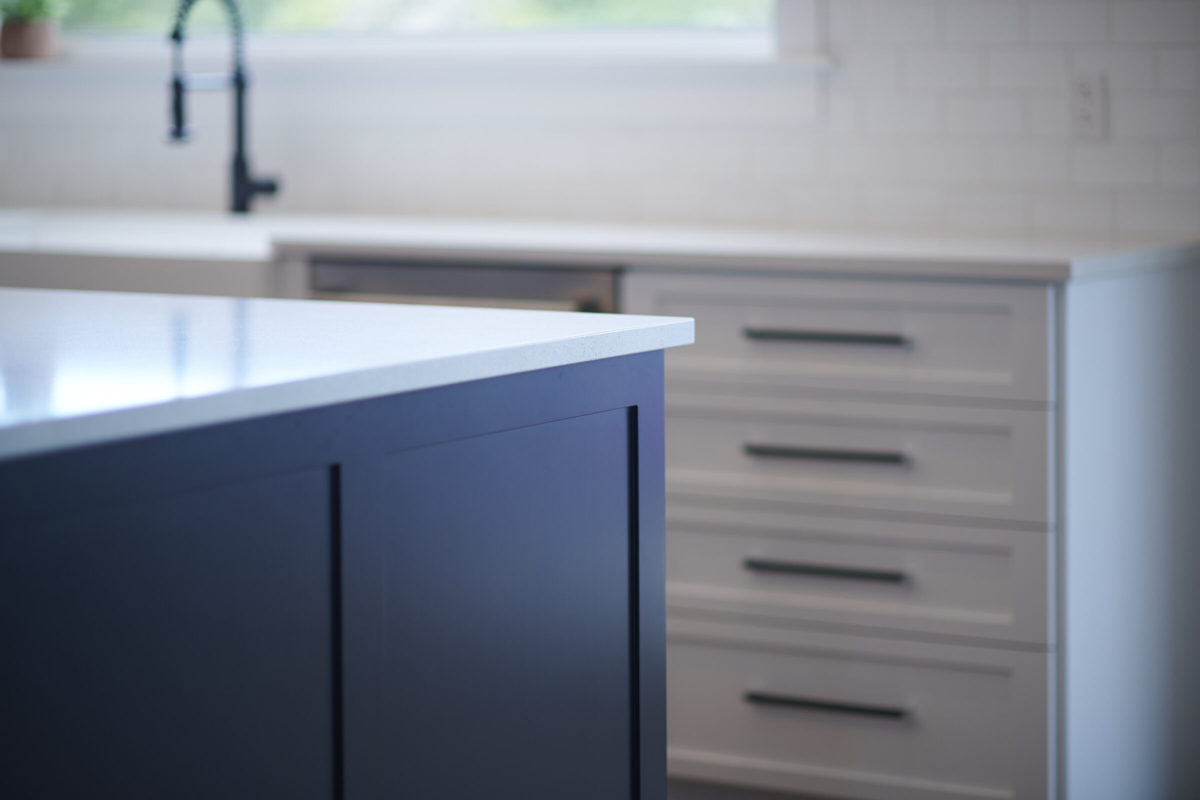 Focused on a dark kitchen island counter with blurred background showing light-colored cabinetry, a sink, and a modern, clean aesthetic.
