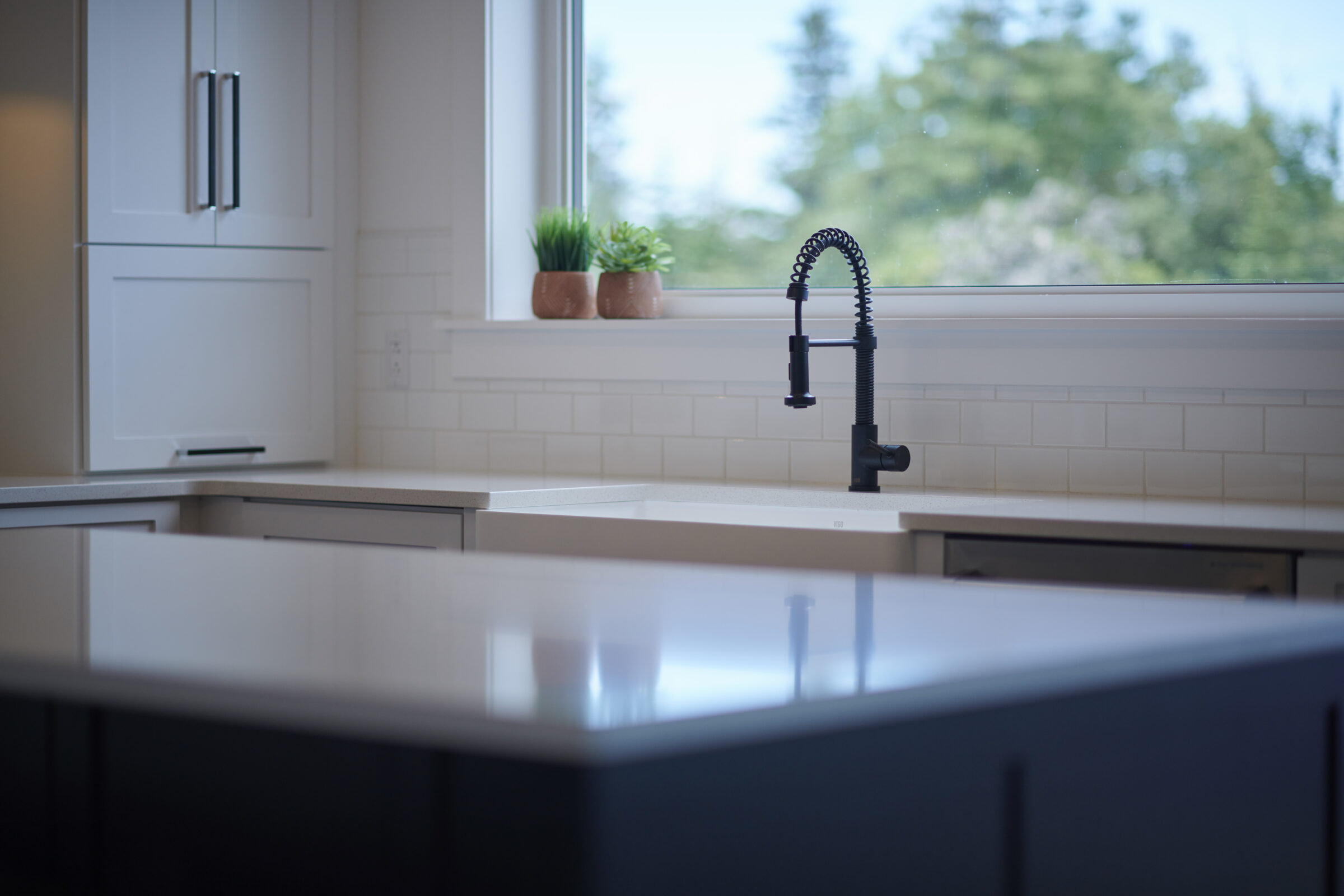 Modern kitchen interior with a reflective countertop, dark sink, stylish faucet, white cabinetry, subway tile backsplash, and green plants by the window.