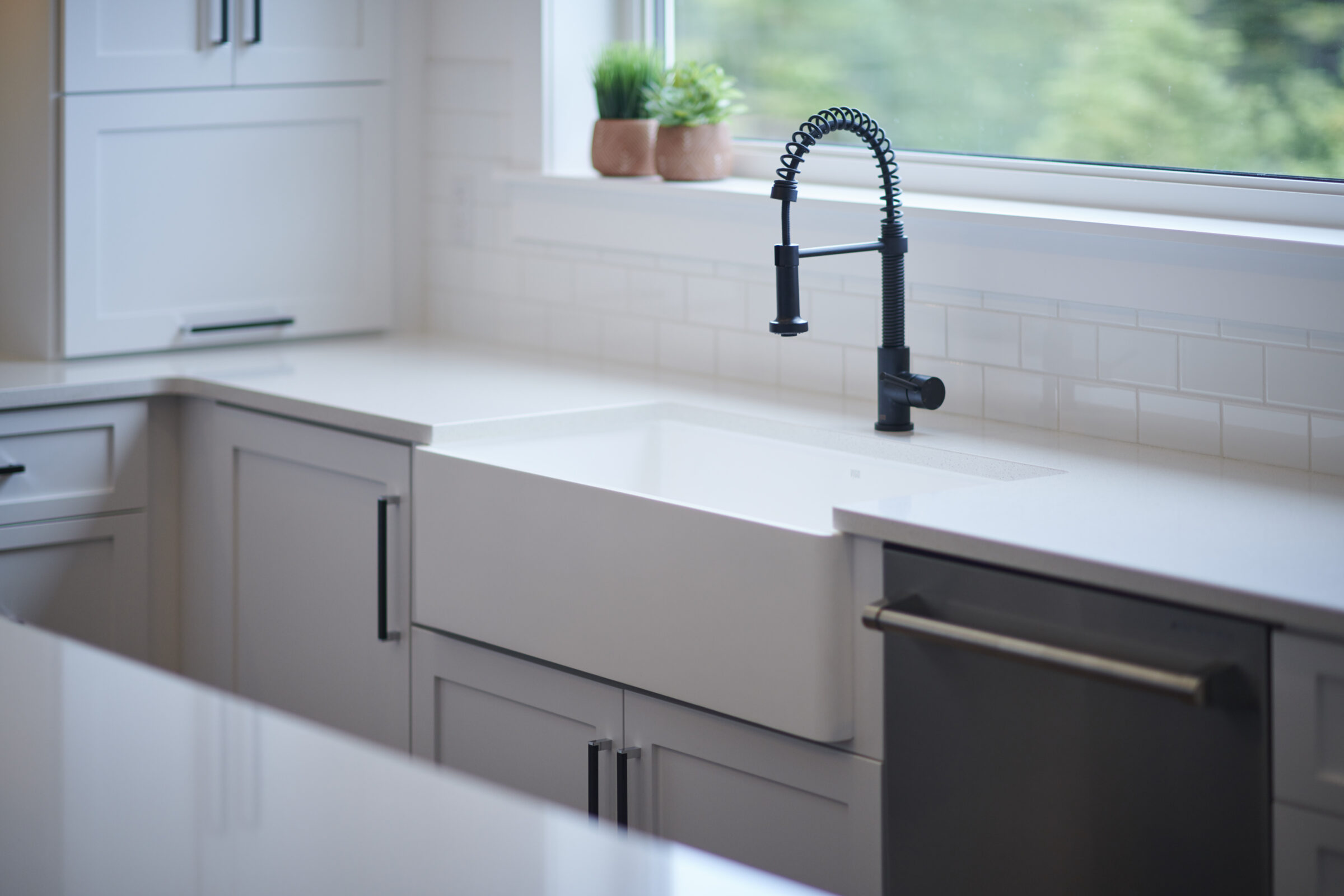 A modern kitchen sink with a black faucet set against a window with a view. White countertops, cabinets, and subway tile backsplash complete the clean design.