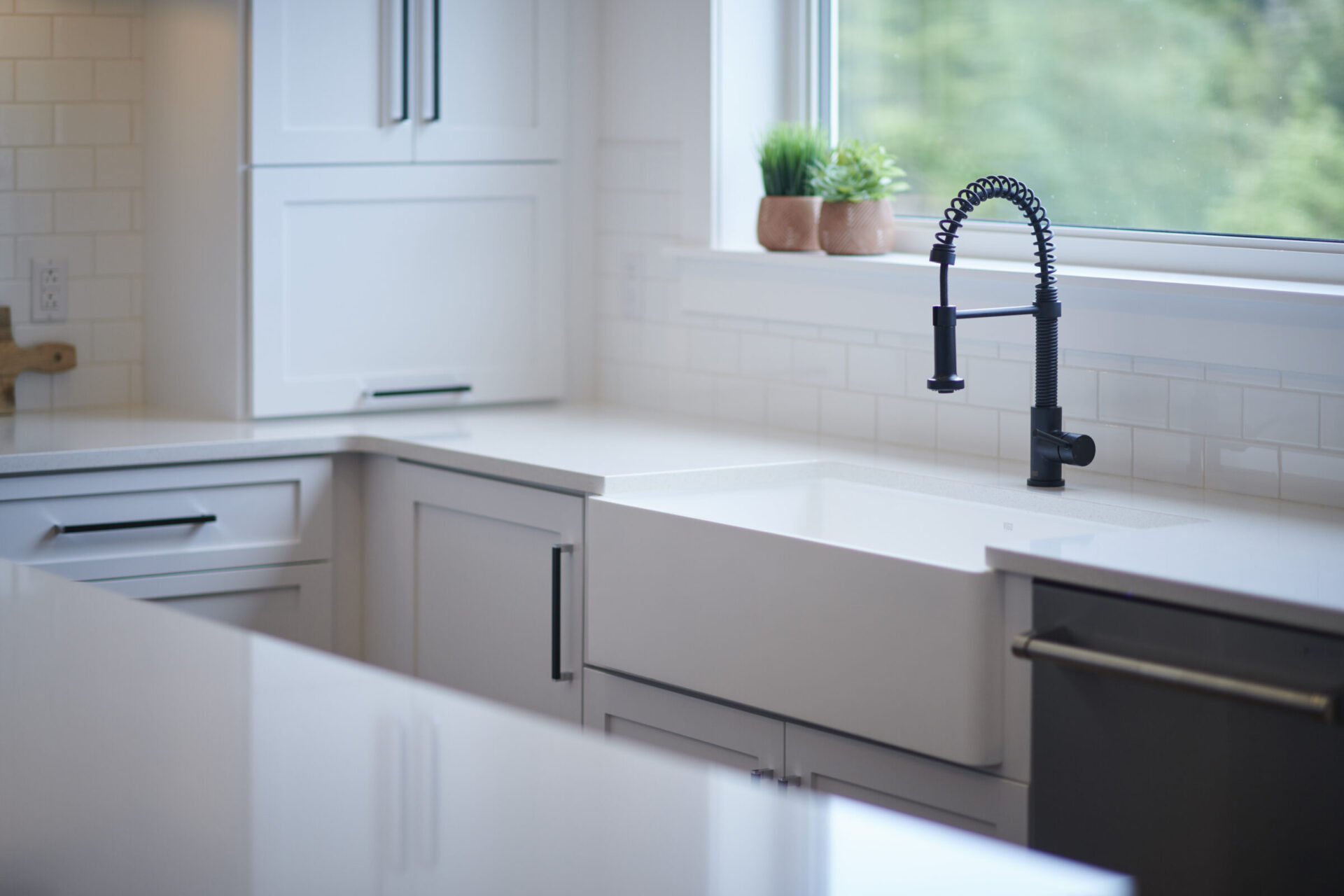 A modern kitchen interior featuring a white countertop, black faucet, subway tile backsplash, white cabinets, and a potted plant by the window.