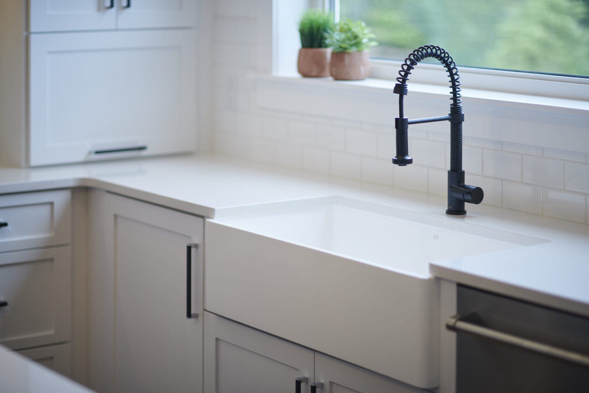 A modern kitchen sink with a black faucet, white countertops, and cabinetry. A window provides natural light and shows greenery outside.