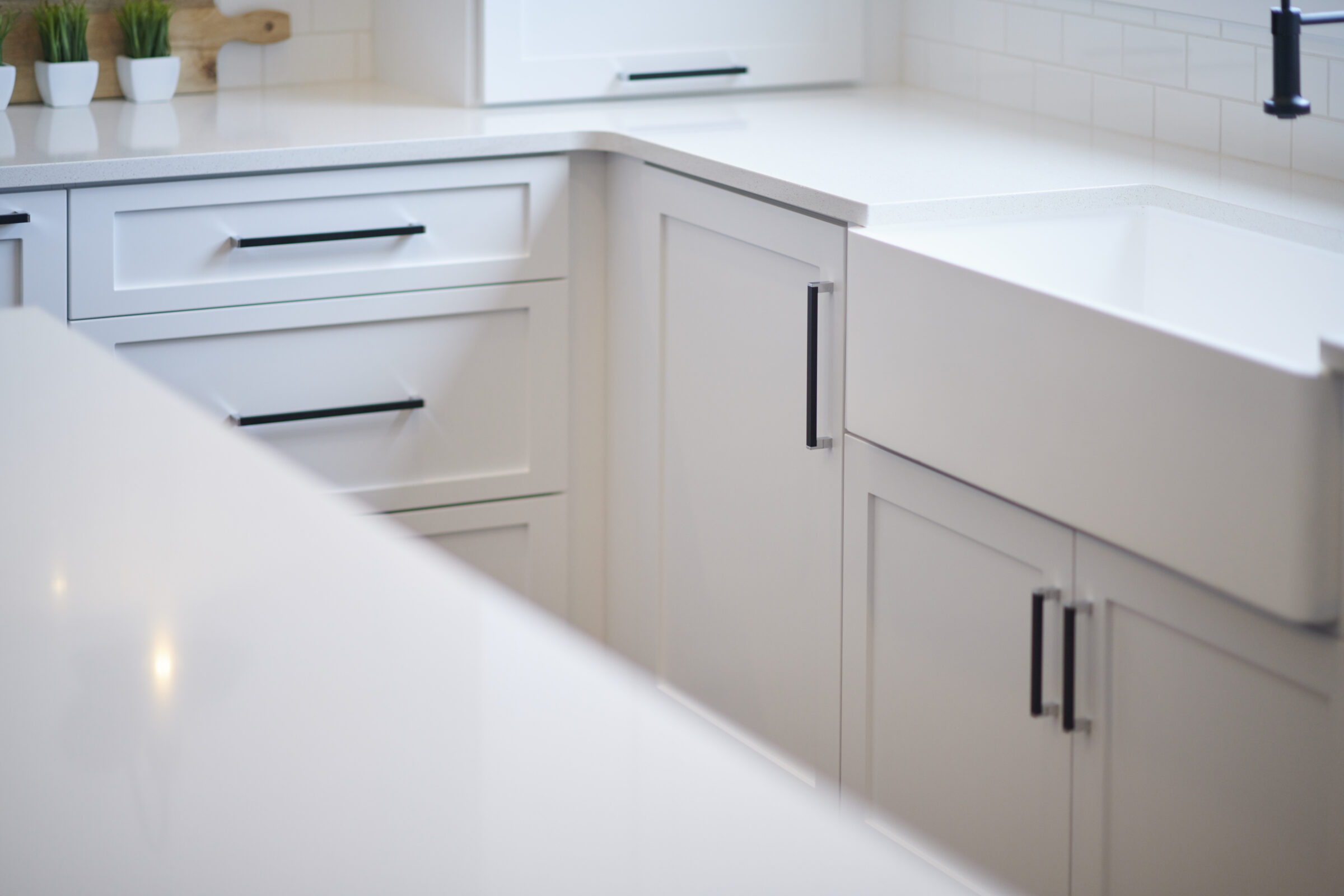 A modern kitchen featuring white cabinets with black handles, a white countertop, and a farmhouse-style sink, with a clean, minimalist design.