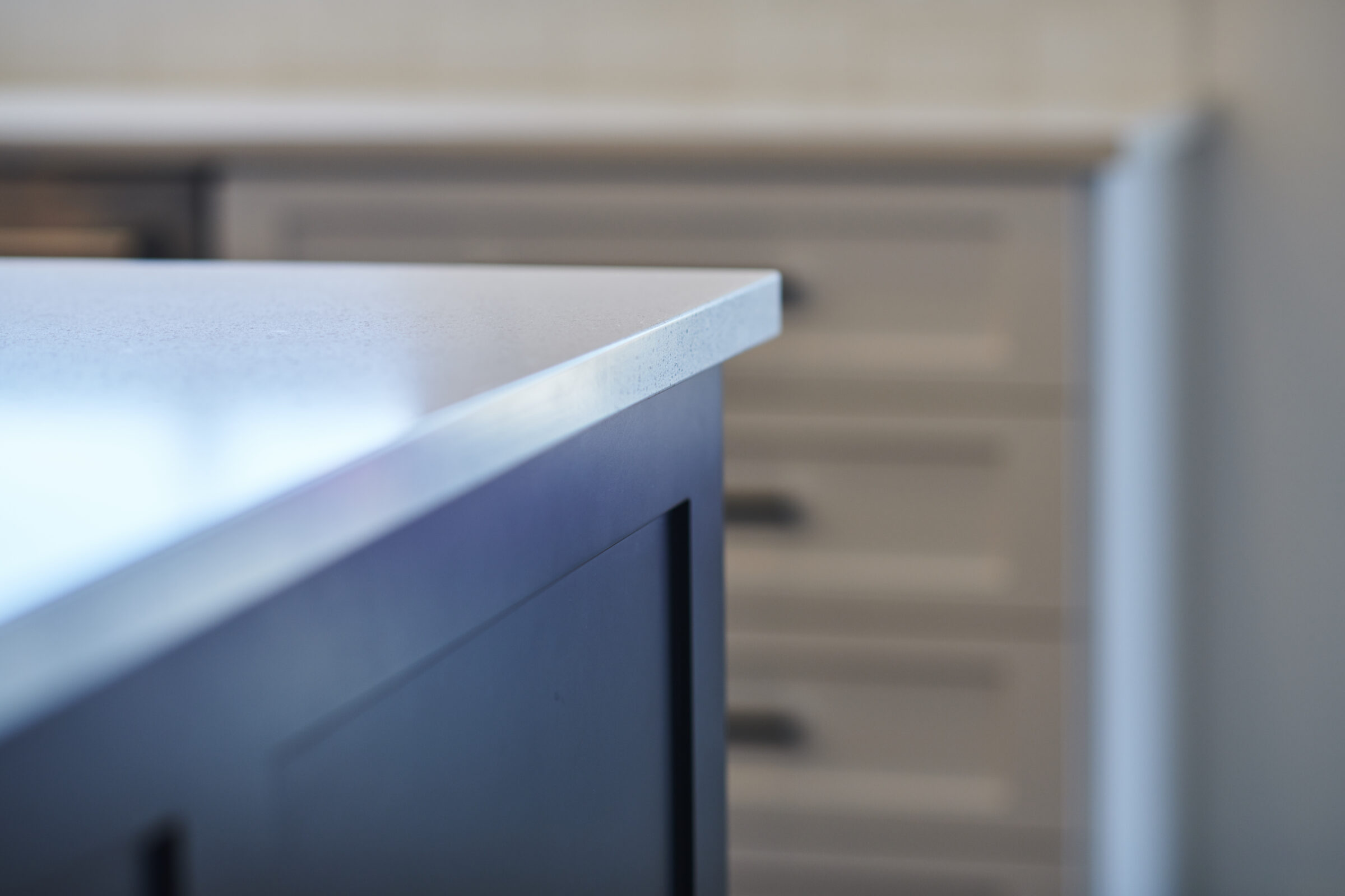 This image shows a close-up view of a kitchen with a focus on a countertop edge, a blurred cabinet in the background, featuring modern materials.