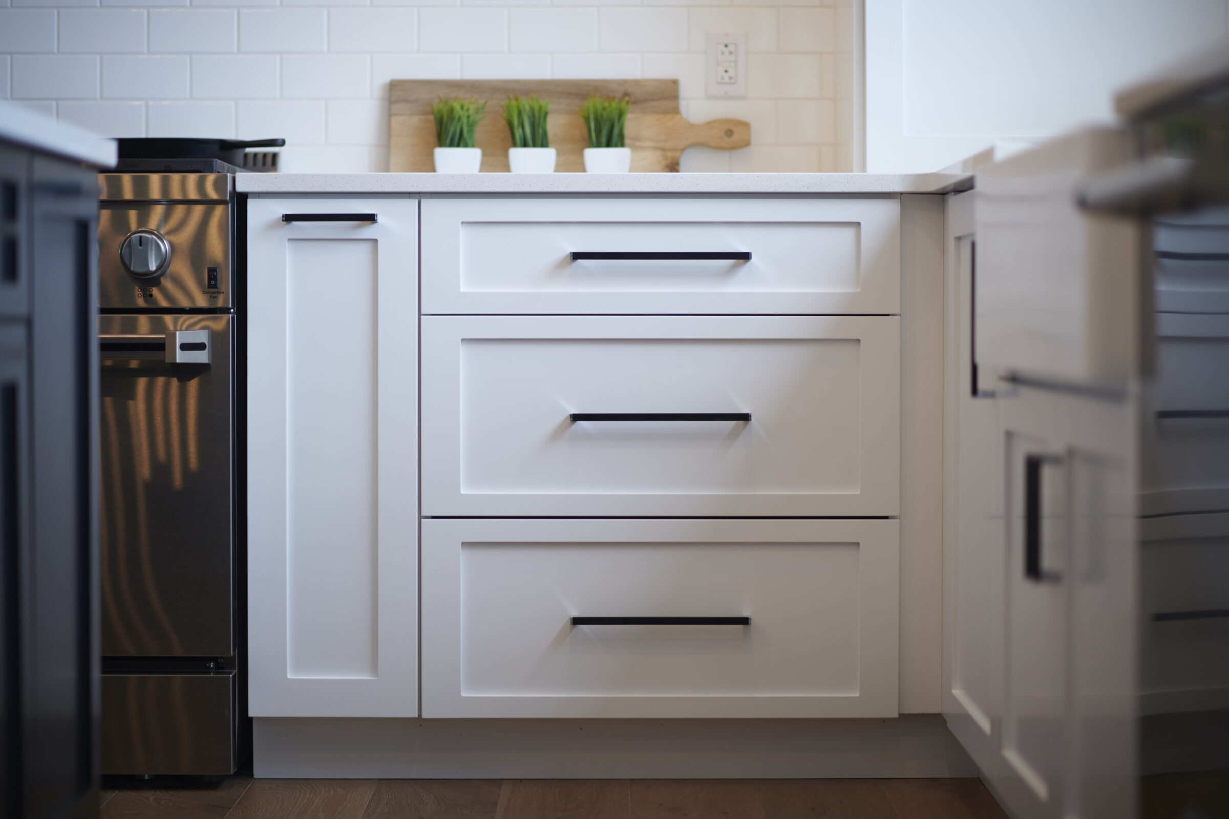 A modern kitchen with white cabinets, stainless steel appliances, subway tile backsplash, and wood accents. Three potted green plants add a touch of nature.