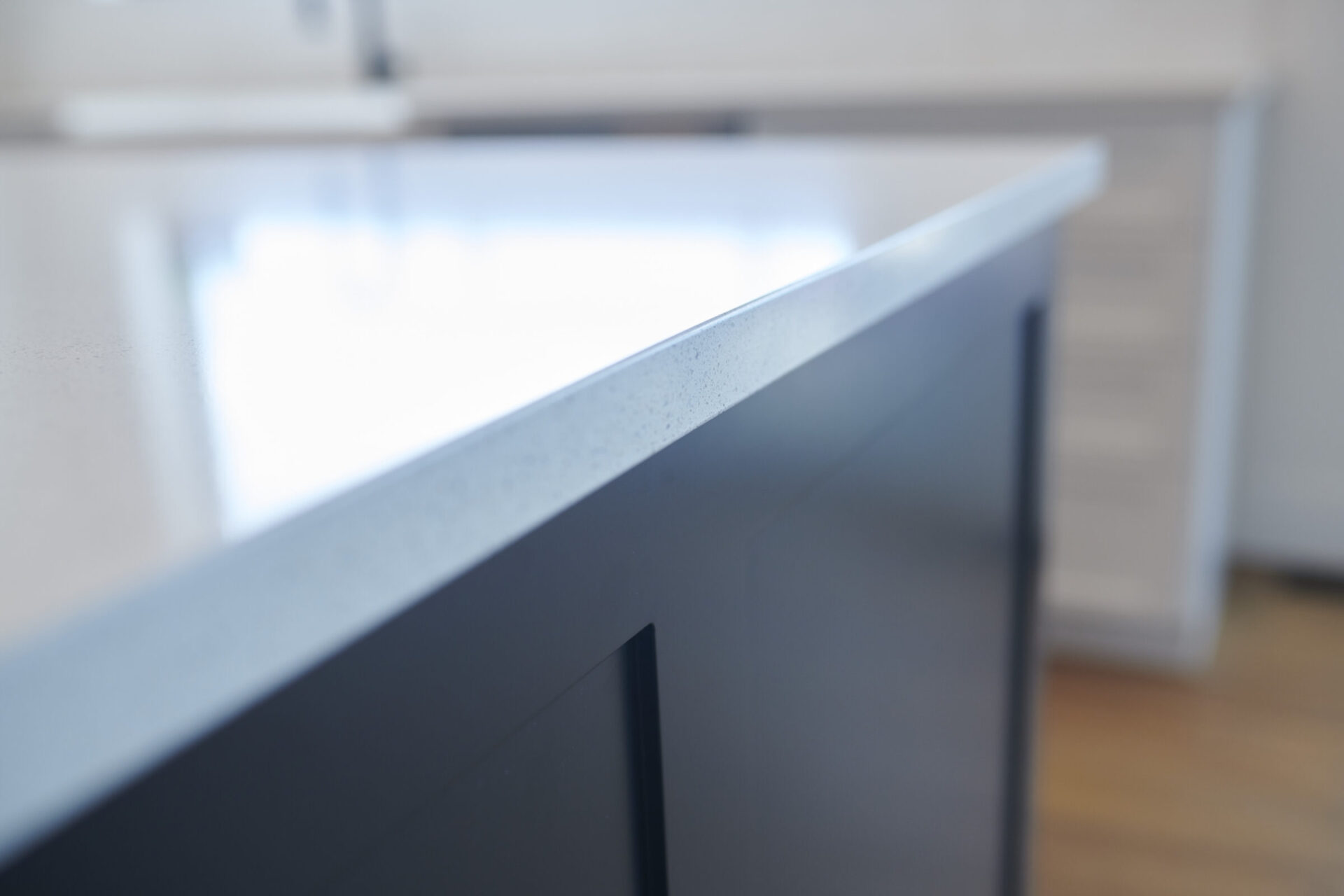 Close-up of a kitchen counter corner showcasing its texture and finish, with blurred cabinetry and flooring in the background, in a minimalist design.