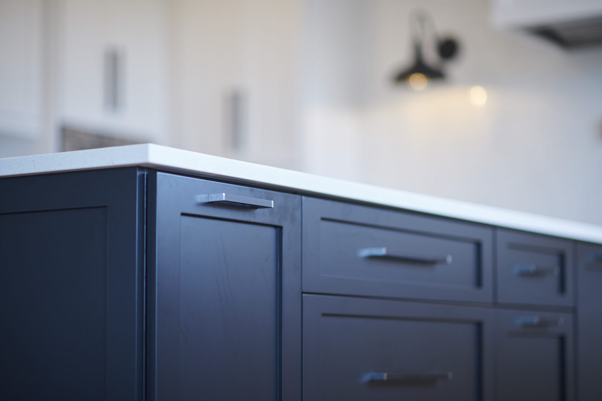 Modern kitchen with dark cabinetry and metallic handles. Out-of-focus background shows a bright wall and part of a wall-mounted light fixture.