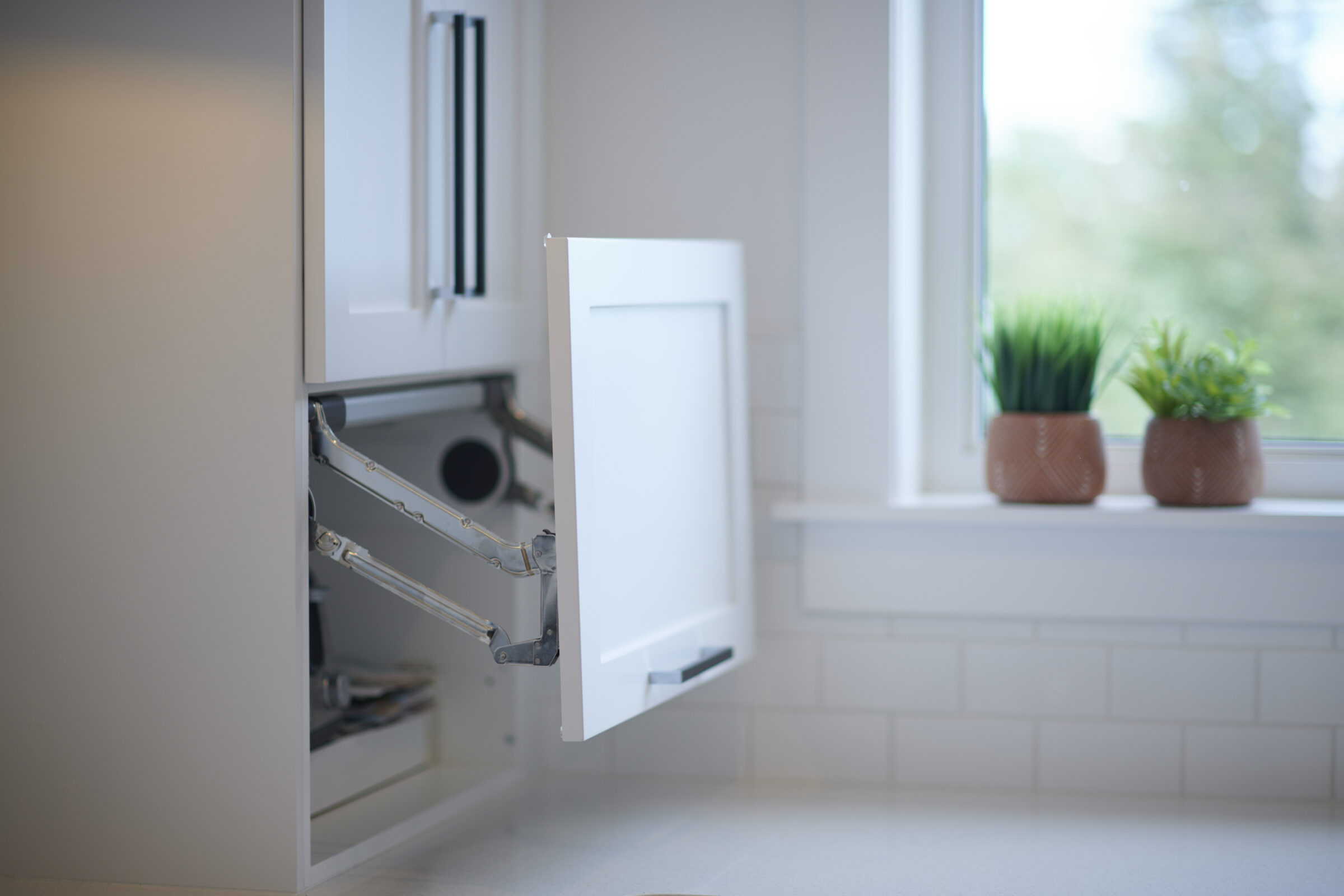 A modern kitchen with an open cupboard revealing waste bins, next to a window with two potted plants on the sill. Clean, bright atmosphere.
