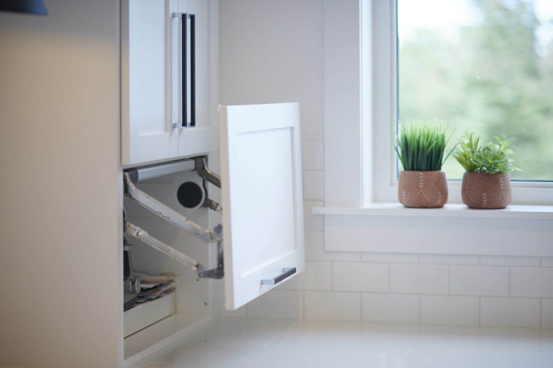 A modern kitchen counter with an open waste bin drawer, next to a window showcasing two potted plants, providing a clean and organized look.
