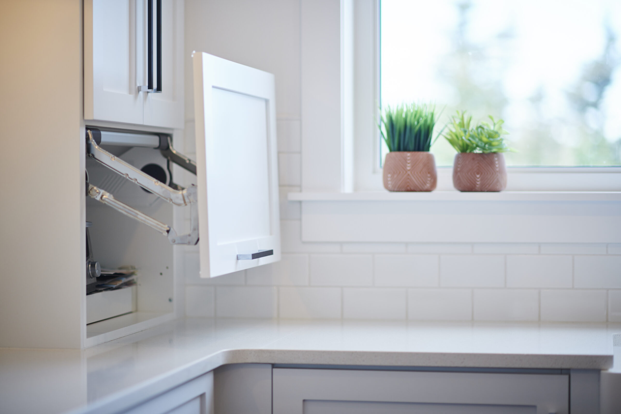 A modern kitchen interior with an open dishwasher, white subway tile backsplash, countertop, and two potted plants on the windowsill with daylight streaming in.