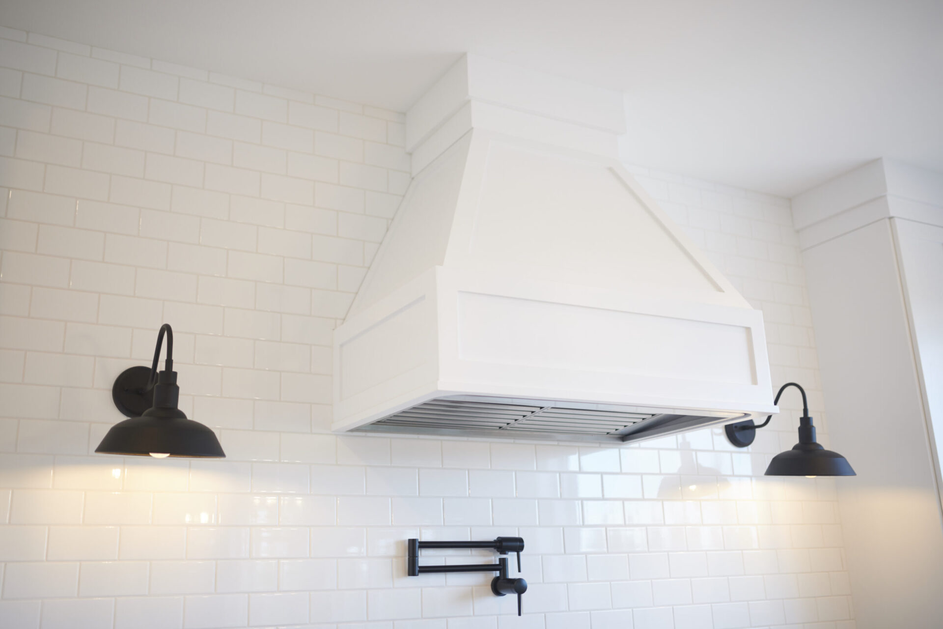 This is a modern kitchen backdrop featuring a large white range hood, subway tile wall, and two black industrial-style wall sconces.