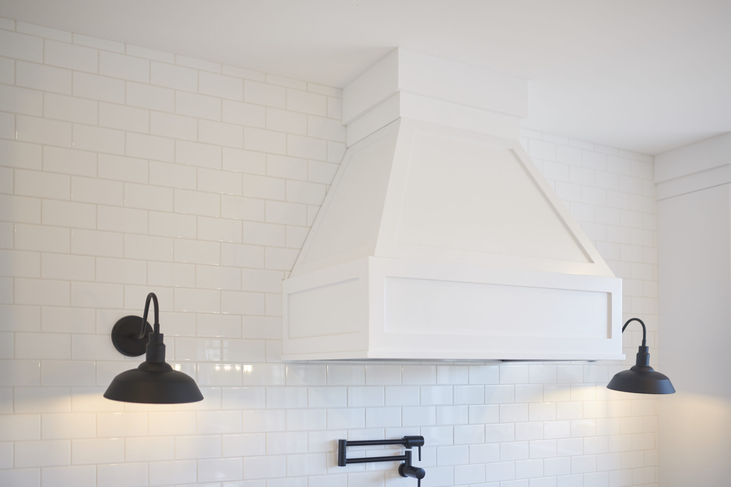 A clean, modern kitchen interior with white subway tiles, a large white range hood, and two black sconce lights illuminating the wall.