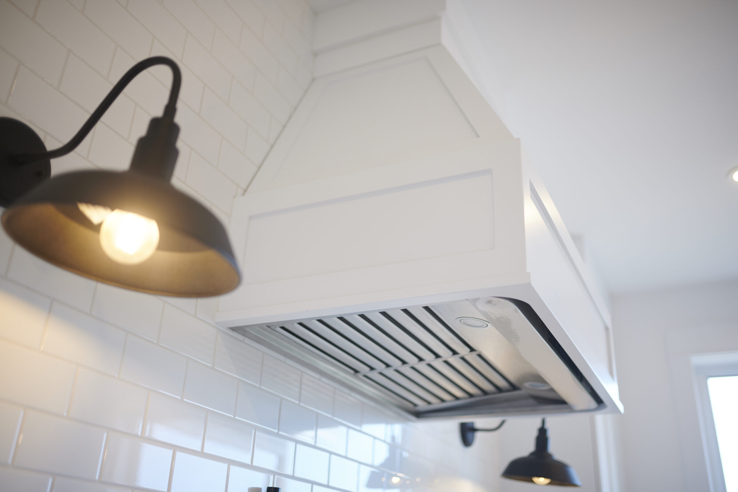 Modern kitchen setting with an illuminated wall-mounted light fixture and a white range hood above a stove, with subway tile backsplash and a bright ambiance.