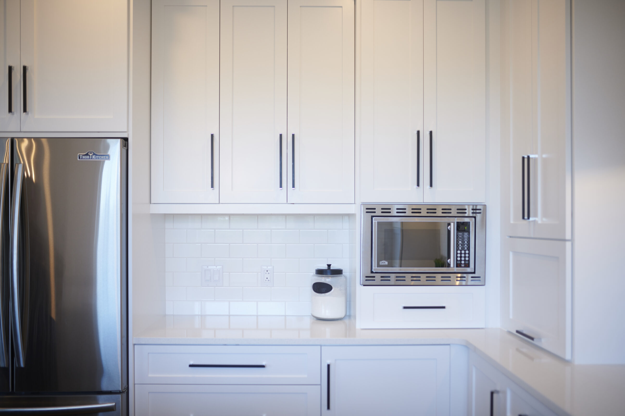 A modern kitchen with white cabinetry, stainless steel refrigerator, built-in microwave, subway tile backsplash, and minimalistic design. Clean and bright interior space.