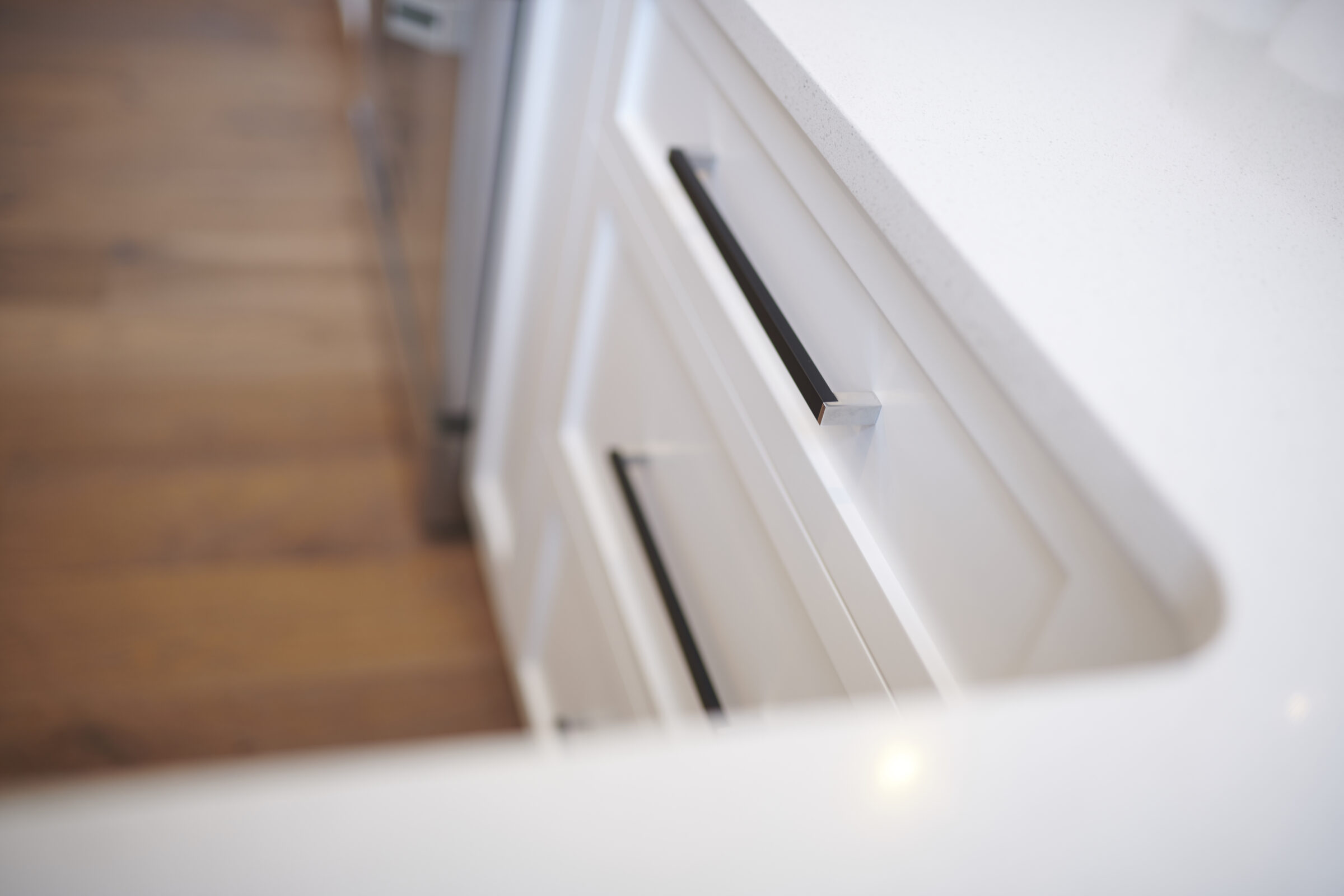 This is a close-up of a white kitchen counter with modern handle design, on wooden flooring. The focus is soft, with a shallow depth of field.