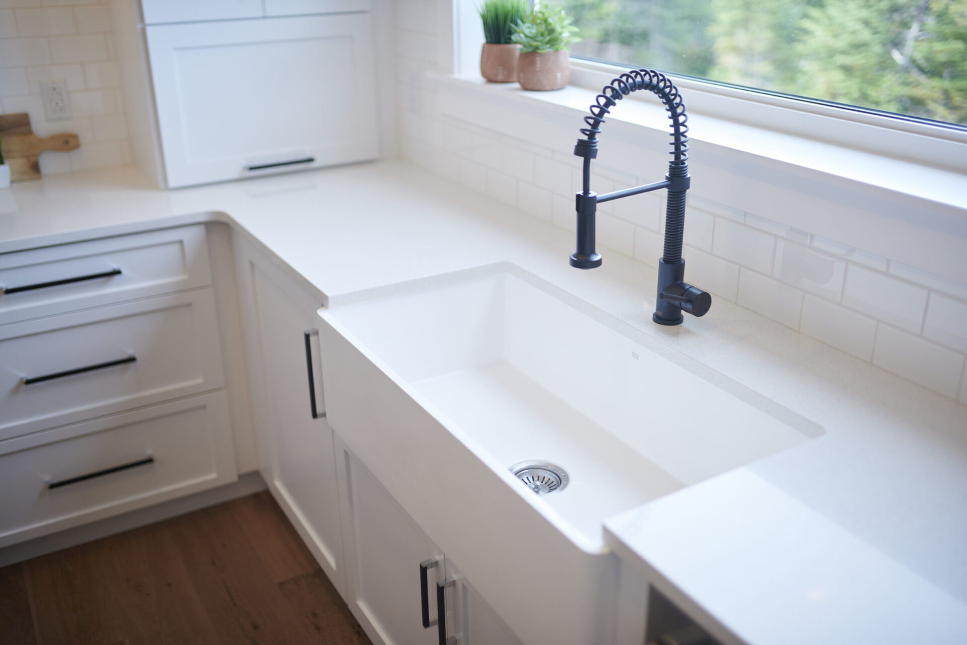 The image shows a modern kitchen corner with a white countertop, a large undermount sink, a black spring faucet, white cabinetry, and a green plant by the window.