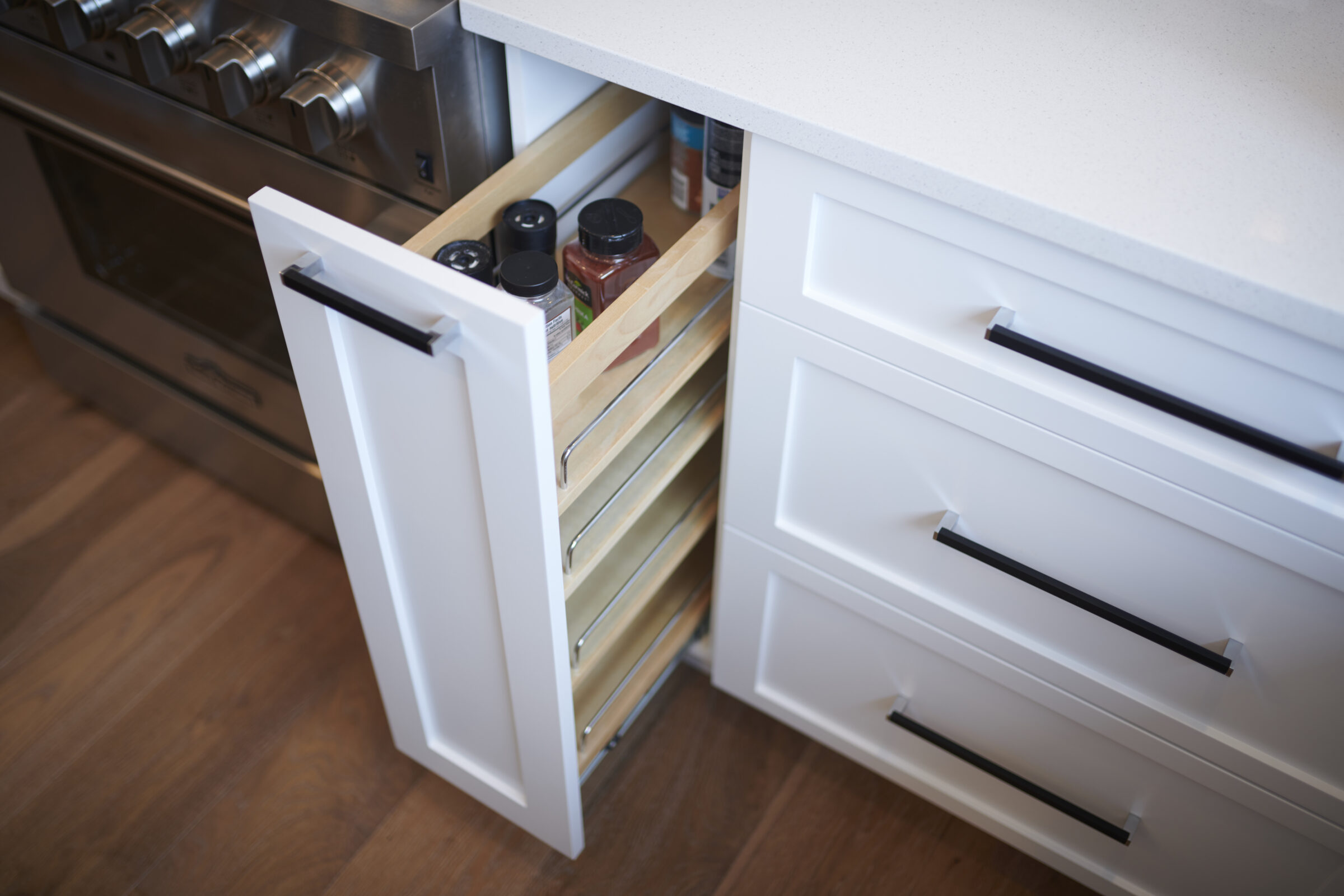 A kitchen drawer is open, revealing neatly organized spice jars. Modern white cabinetry with black handles complements the contemporary stainless-steel oven to the left.
