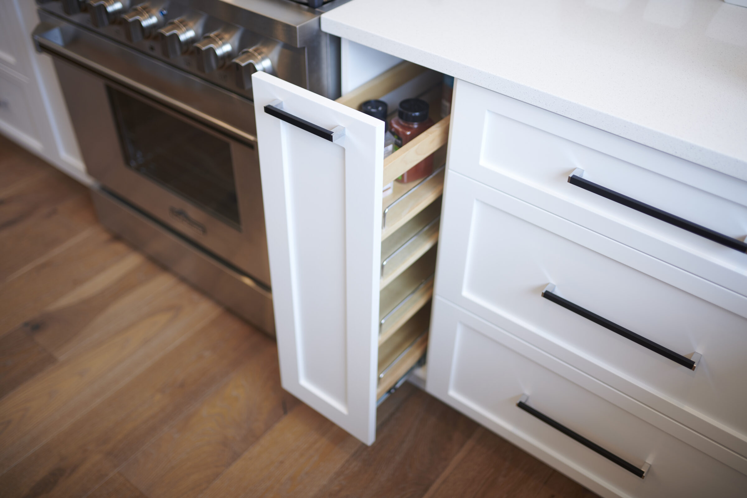 Modern kitchen with stainless steel stove and white cabinets. A drawer is partially open, revealing a wooden spice rack with bottles inside.