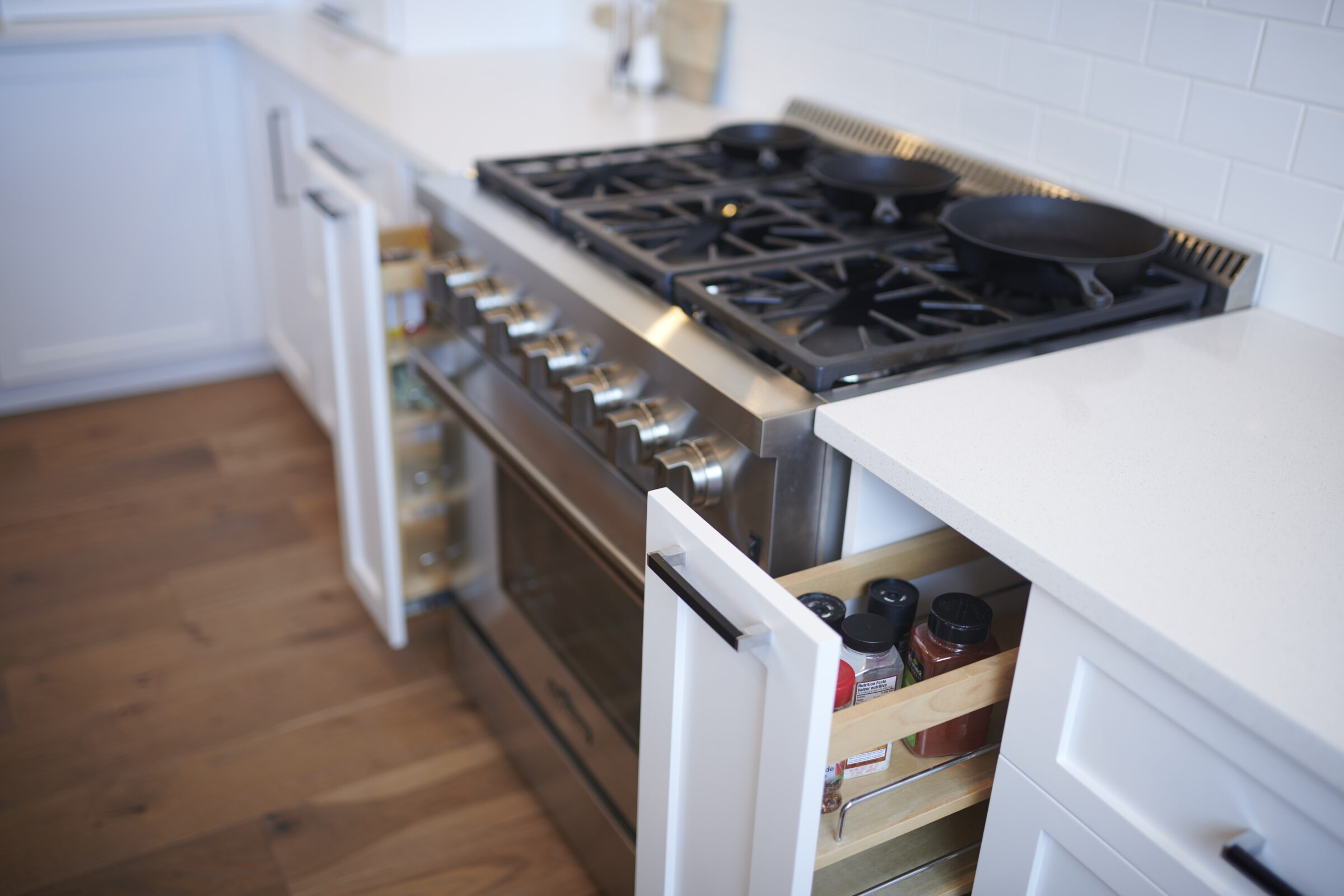 Modern kitchen with stainless steel stove and gas cooktop. Wood flooring, white cabinets, open spice drawer, and cast iron pans are visible.