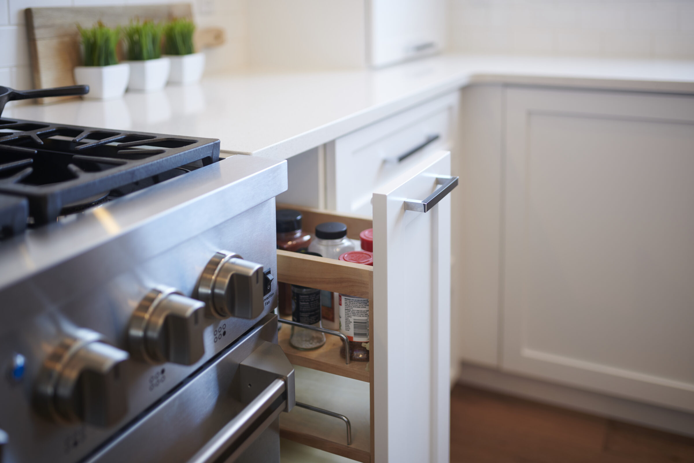 A modern kitchen with a stainless steel gas stove next to an open drawer containing spices and ingredients; pots of herbs in the background.