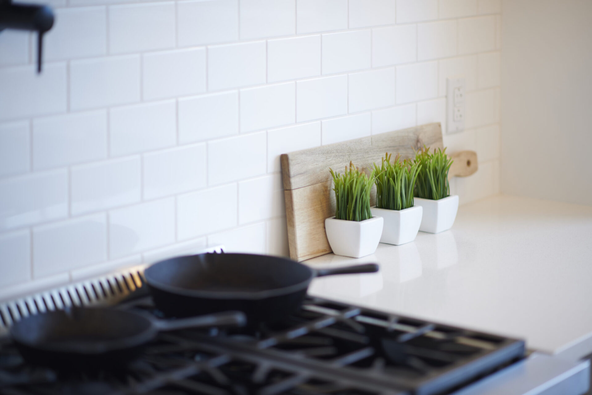 A modern kitchen counter featuring a gas stove, two cast iron pans, and three white pots with green plants against a white subway tile backsplash.