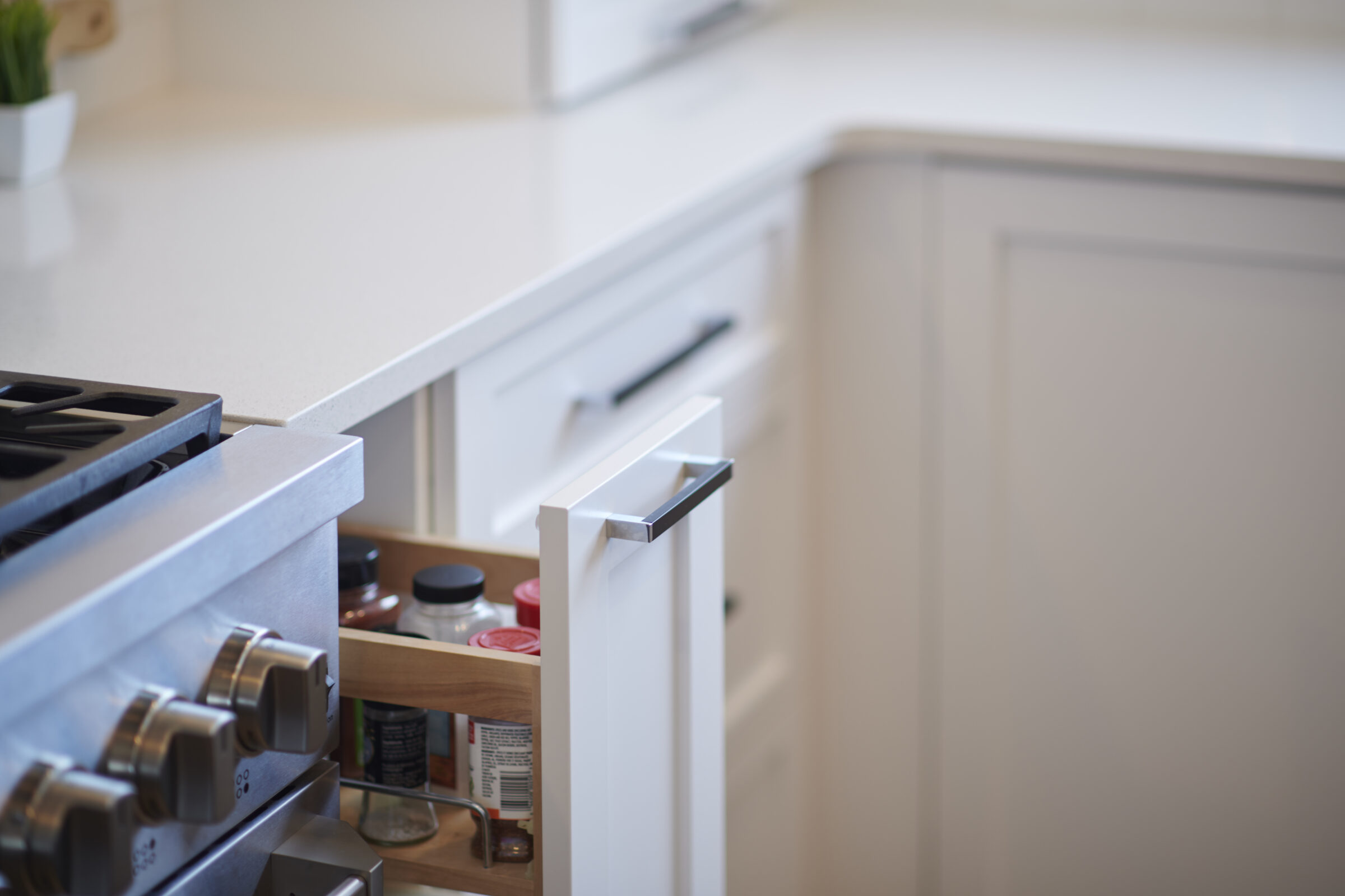 A modern kitchen with white countertops, a gas stove, and an open drawer revealing spices and condiments in a neat arrangement.