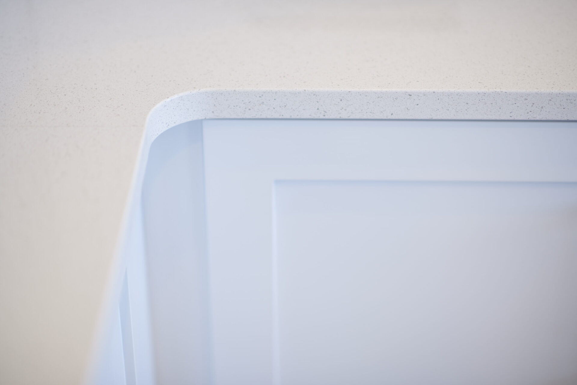 The image shows a close-up of a white kitchen cabinet with a minimalist design and a speckled white countertop, softly focused with a light background.