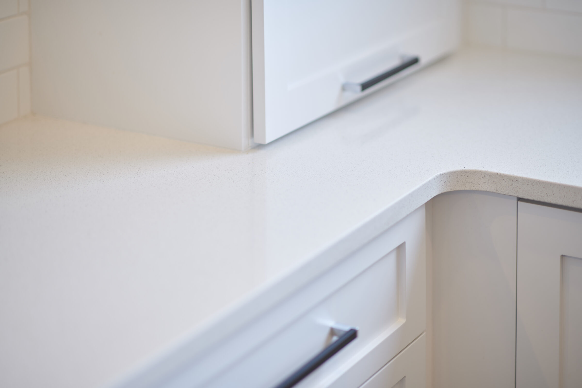 A modern kitchen counter with a white speckled surface, sleek cabinet drawers, and a minimalistic handle. The background features clean subway tiles.