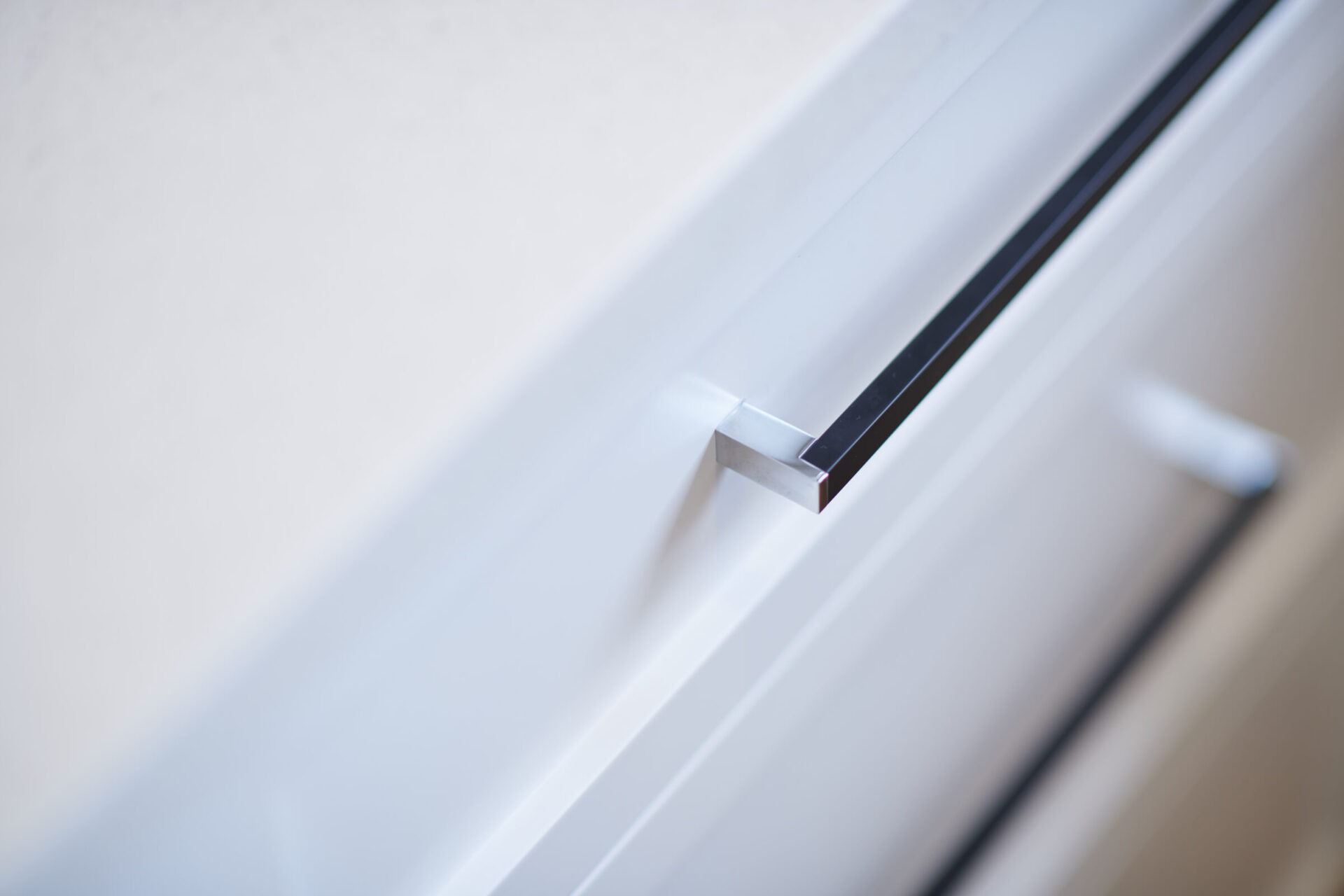 This image shows a close-up of a modern, sleek cabinet handle attached to a white cupboard, with a blurred background emphasizing the handle's clean lines.