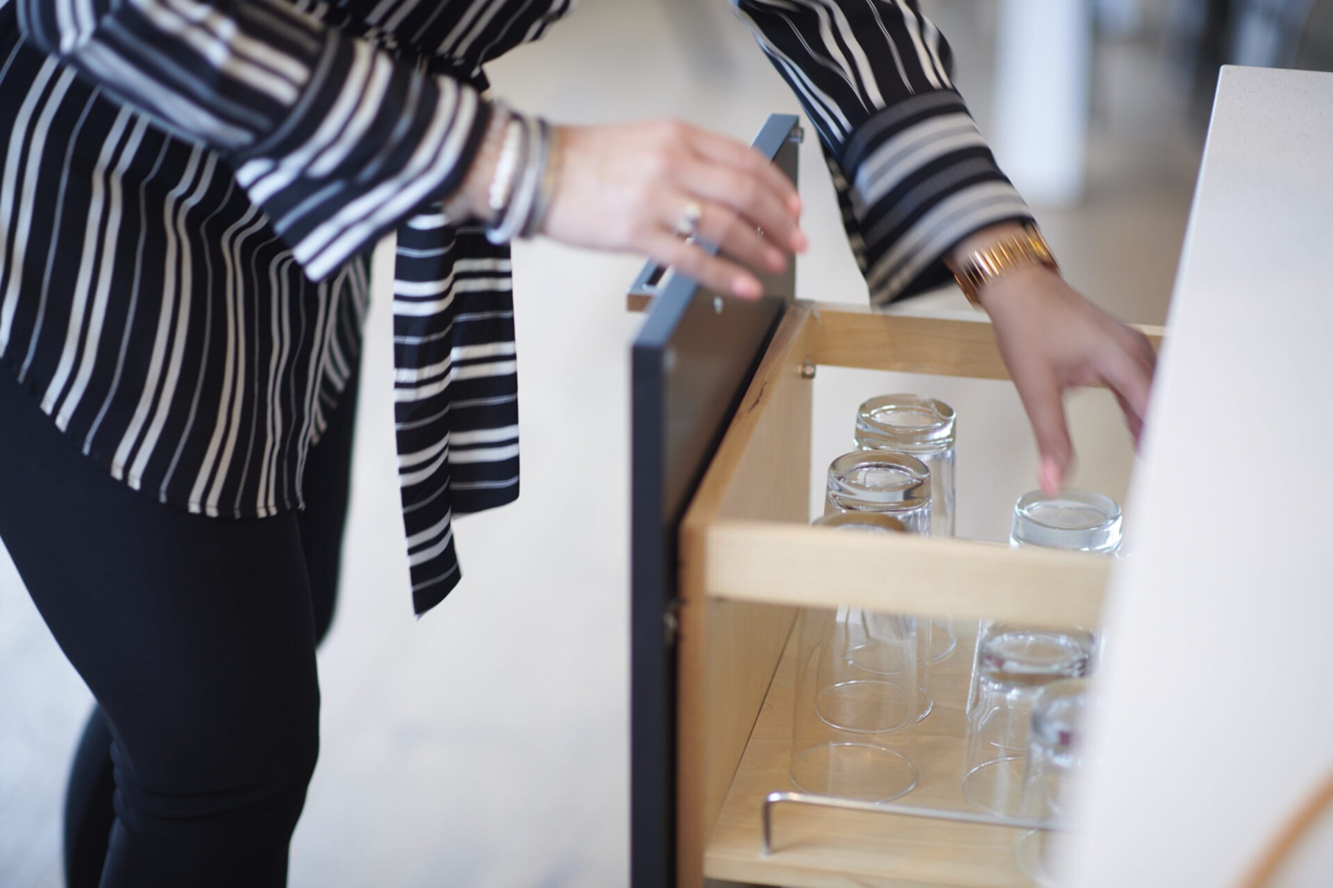 A person in a striped blouse opens a wooden drawer containing several clear glass jars on a light interior background.