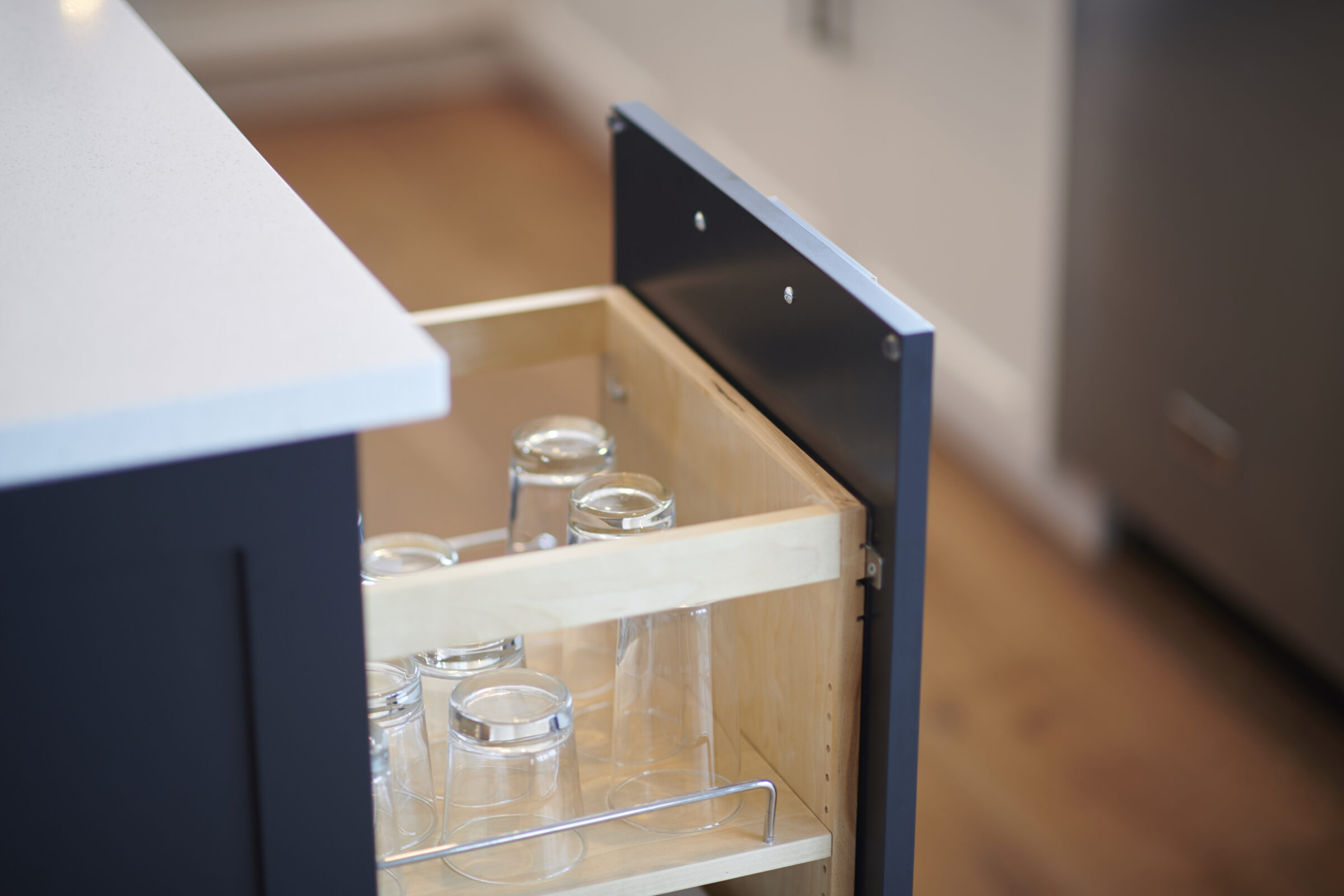 An open kitchen drawer with a variety of transparent glass jars and containers inside, set against wooden cabinetry and a white countertop.