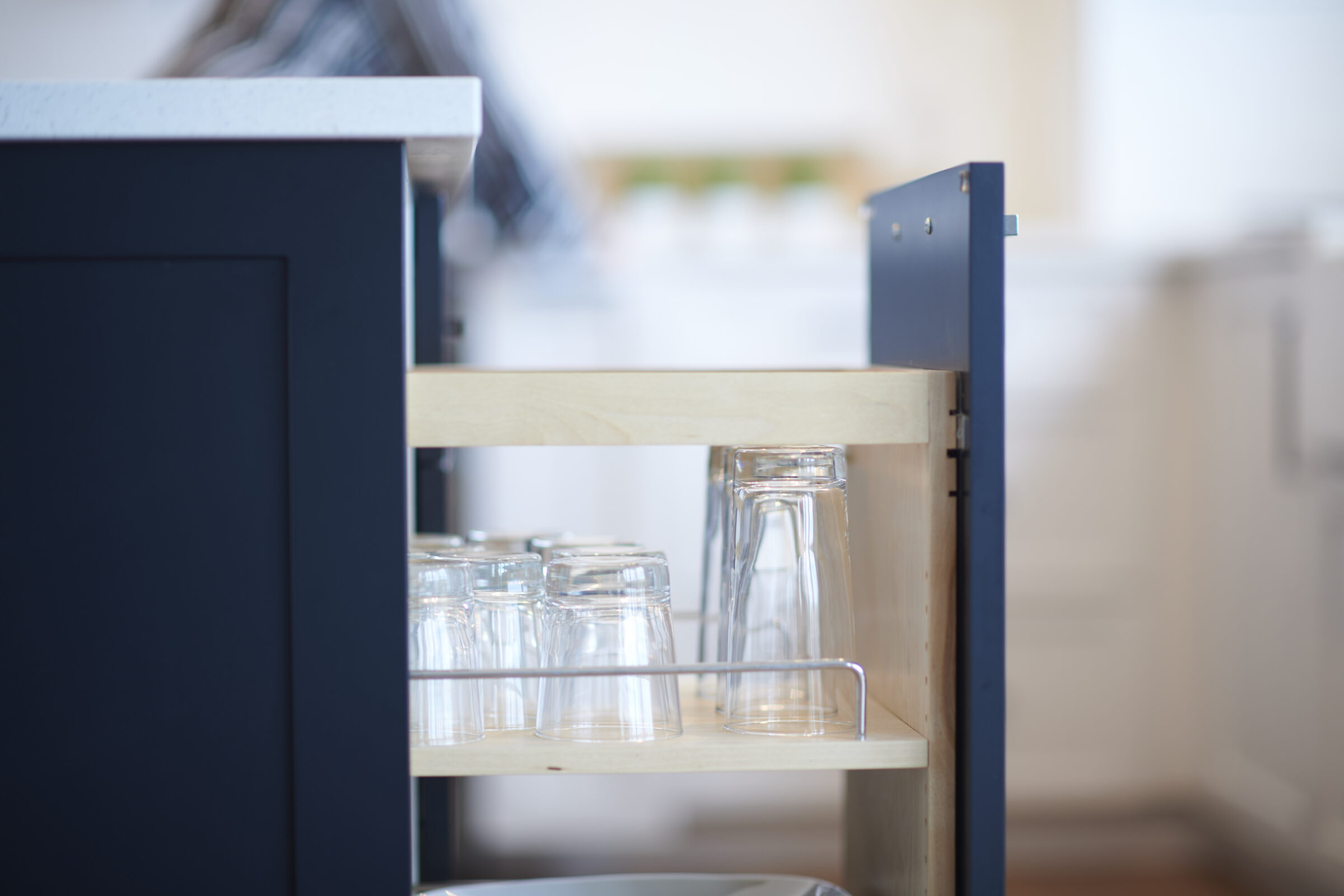 A shallow depth of field focuses on clear glasses neatly arranged on wooden shelves of a cart, with blurred kitchen interiors in the background.