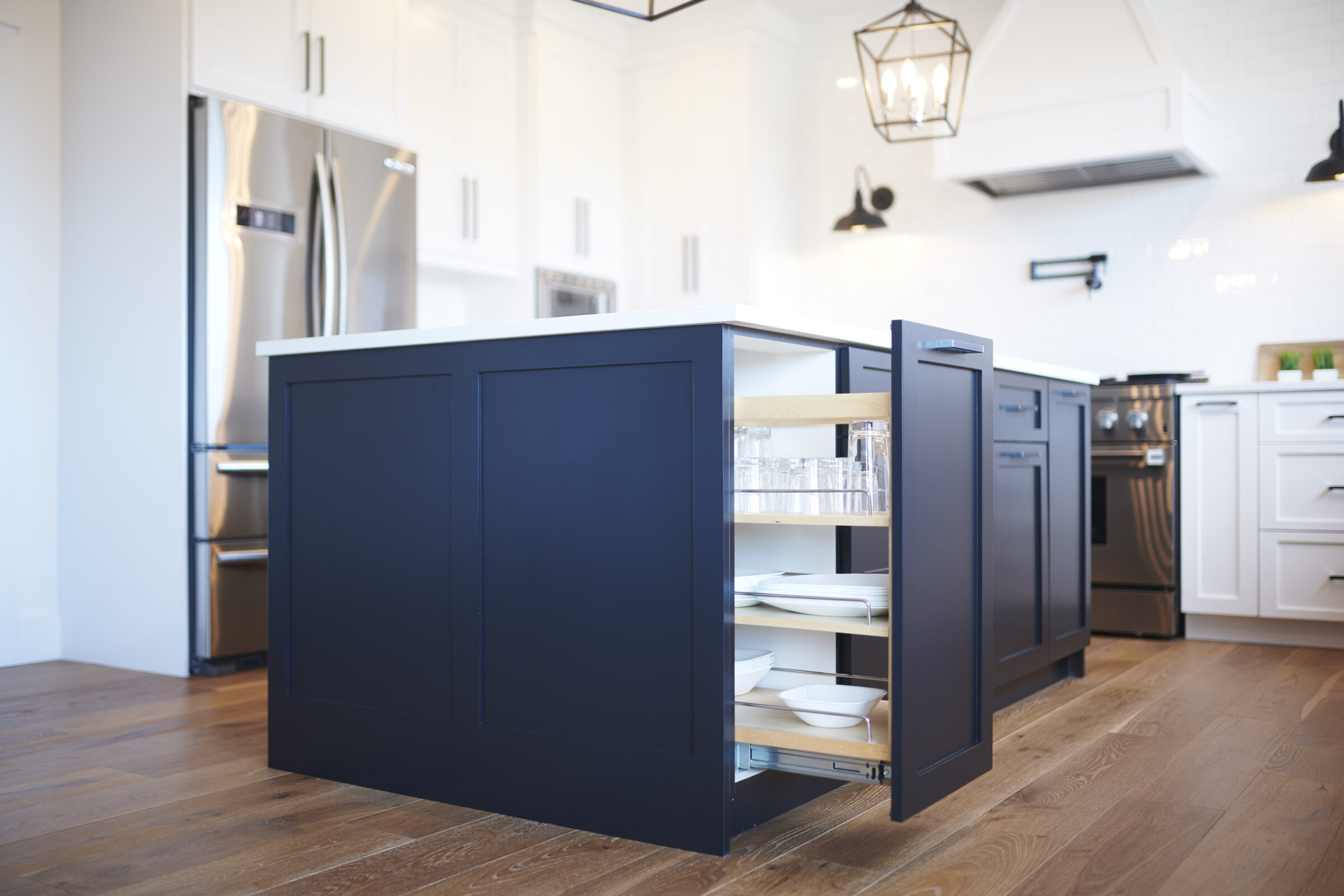 A modern kitchen with white cabinets and a dark blue island featuring open drawers revealing organized dishes and glassware on shelves.