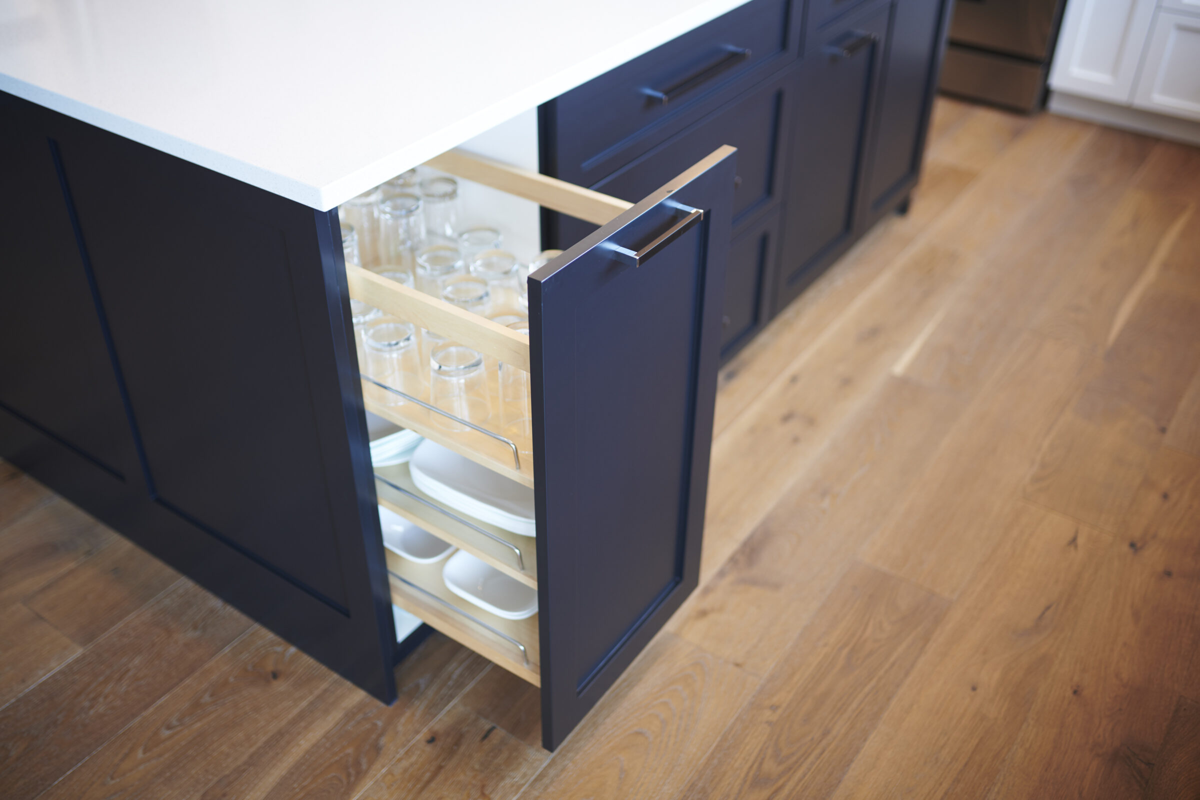 A modern kitchen cabinet drawer is open, revealing organized glassware and plates on two shelves, with a wooden floor and darker cabinetry design.