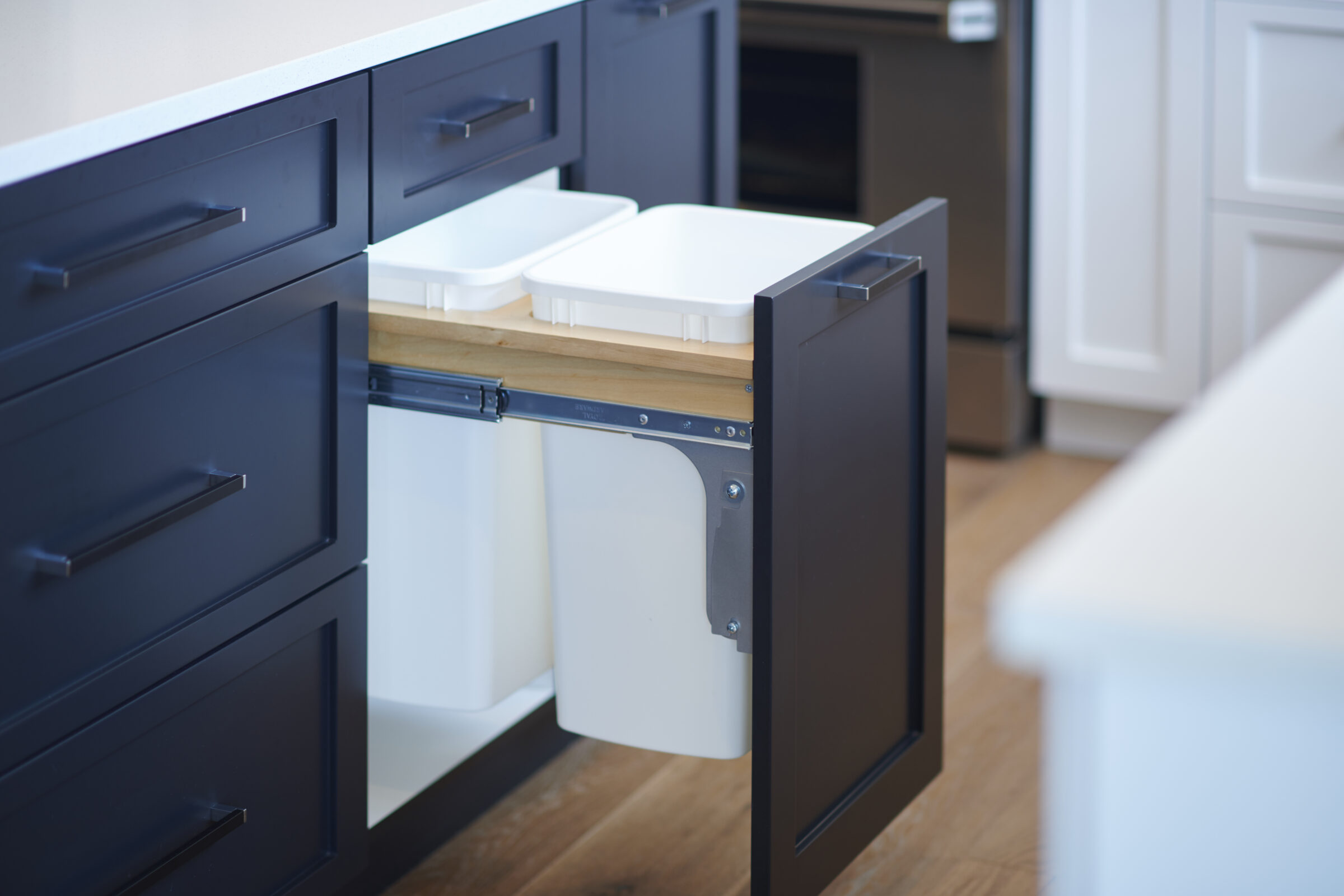 The image shows a modern kitchen with a pull-out waste container system installed in a dark blue cabinet, featuring two white bins with lids.