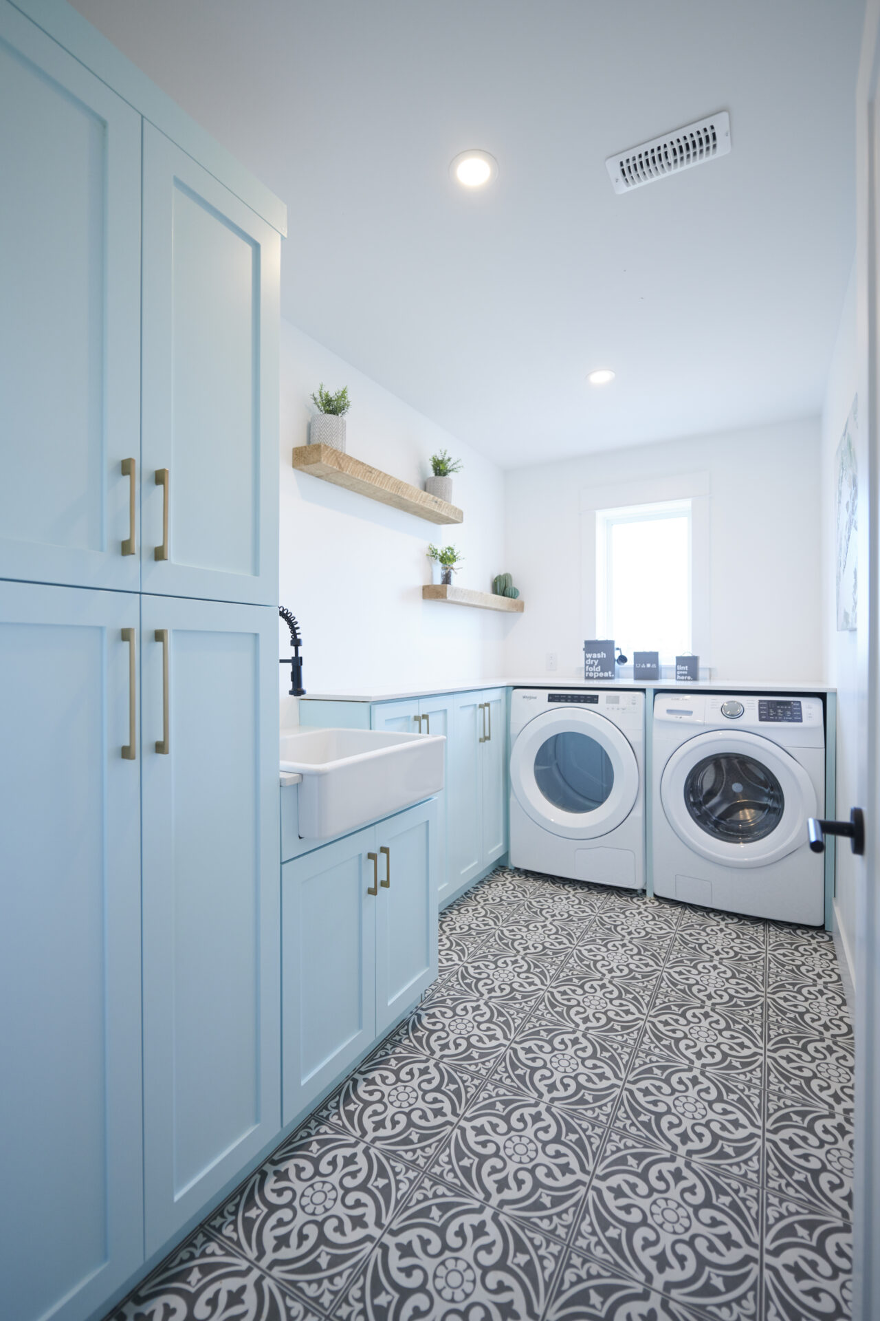 A bright laundry room with blue cabinetry, patterned floor tiles, a farmhouse sink, front-loading washer and dryer, and wooden shelves with plants.