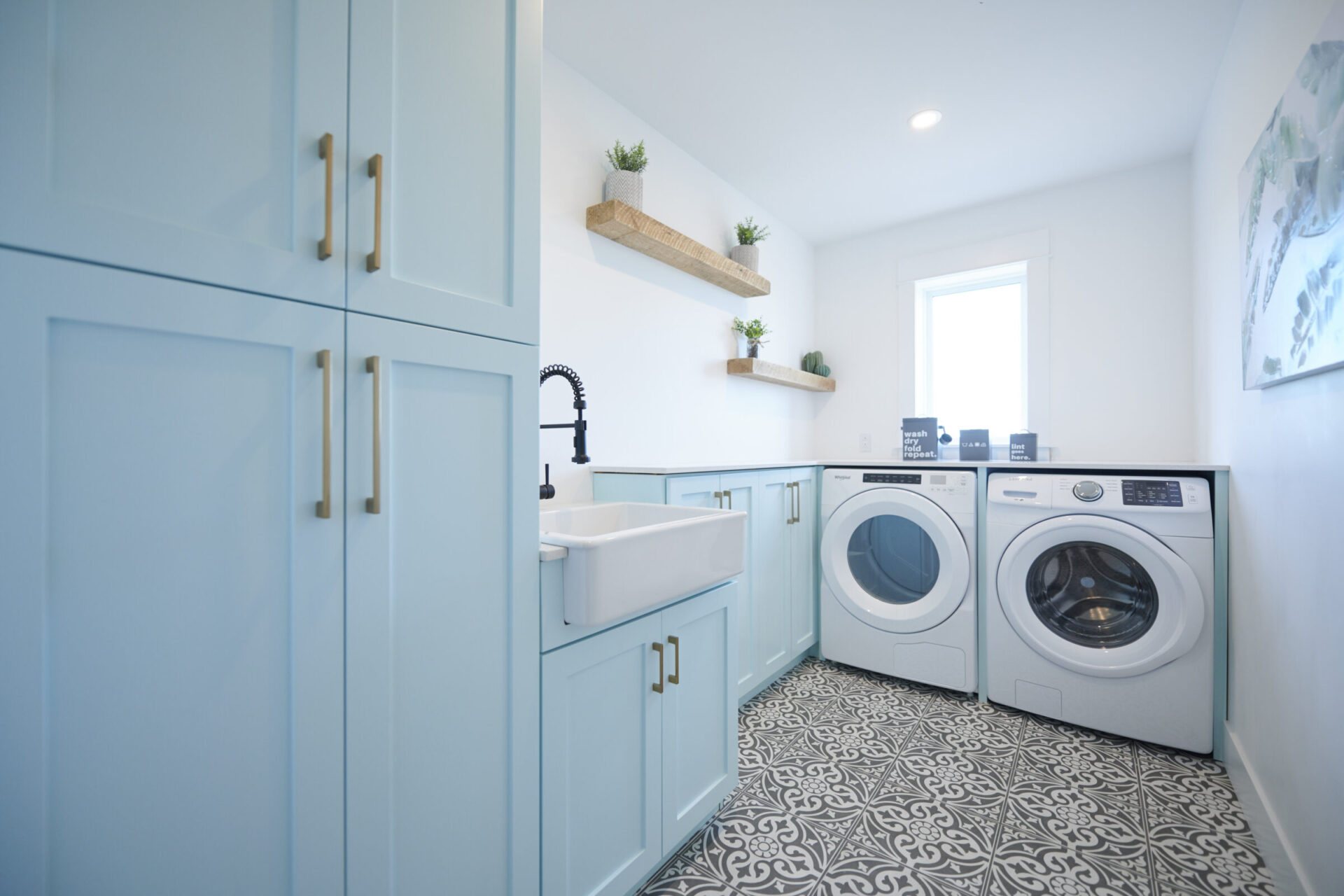 This image shows a bright laundry room with blue cabinets, a white utility sink, front-loading washer and dryer, and decorative shelving with plants.