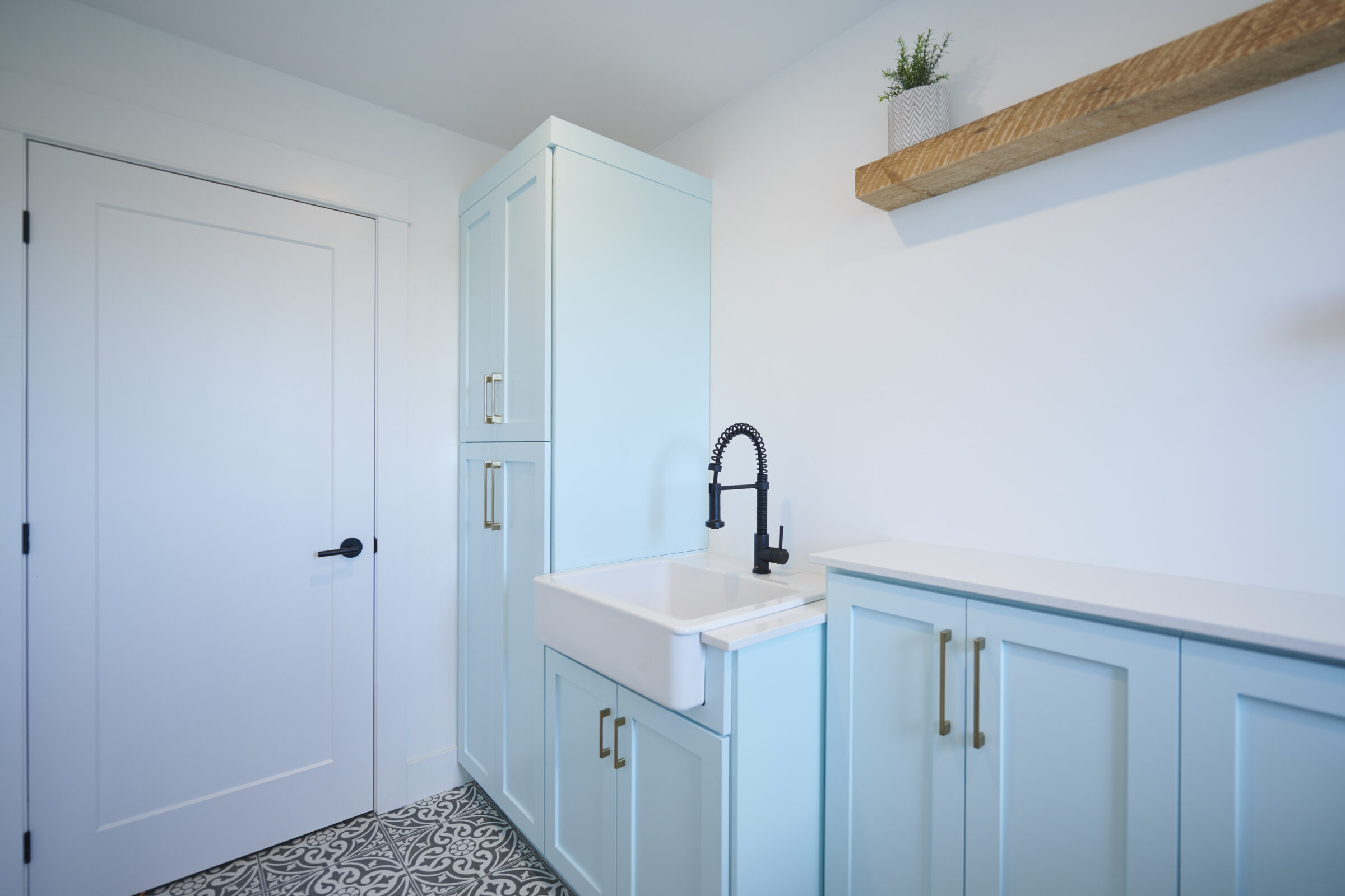 This is a bright, minimalist laundry room with light blue cabinets, a white farmhouse sink, black faucet, patterned floor, and a floating wooden shelf.