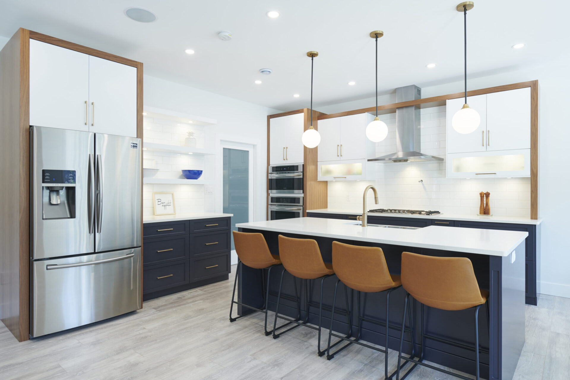 A modern kitchen interior with stainless steel appliances, white countertops, dark cabinets, and a central island with mustard-colored bar stools under pendant lights.