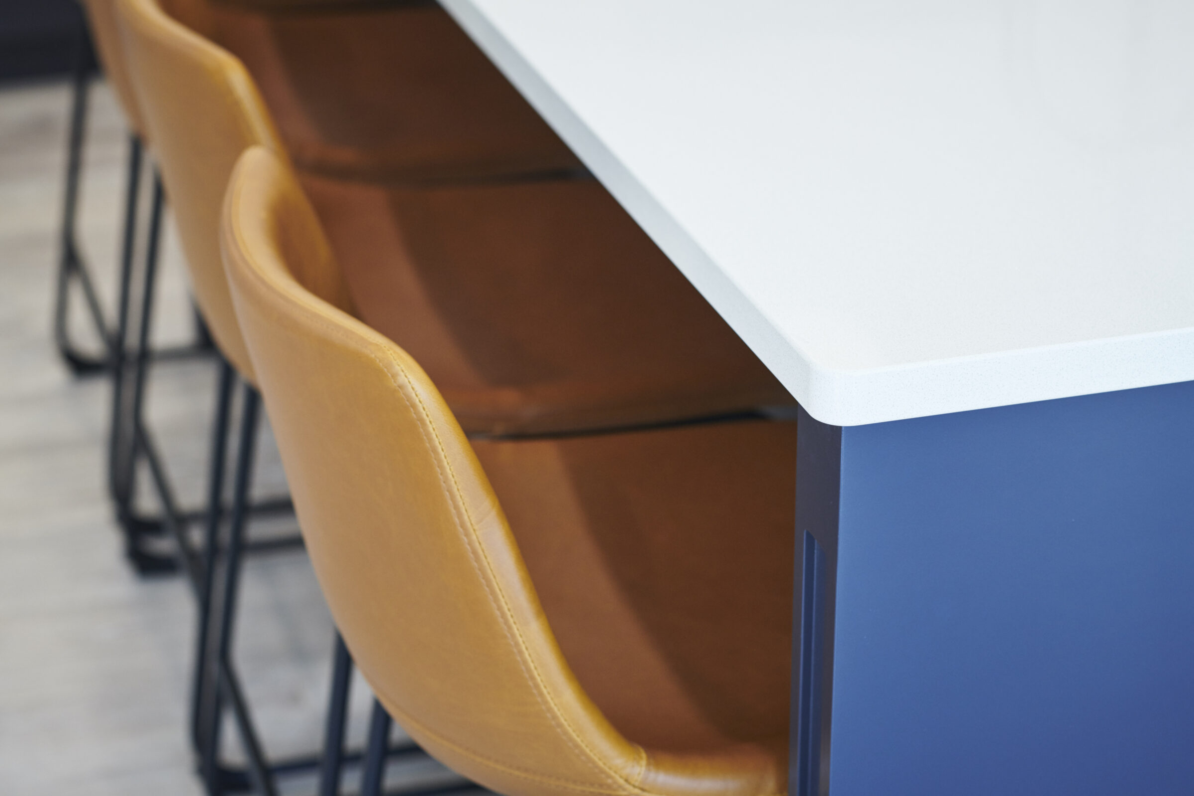 A modern kitchen interior with a white countertop, paired with tan leather bar stools and a blue cabinet, set against a grey floor.