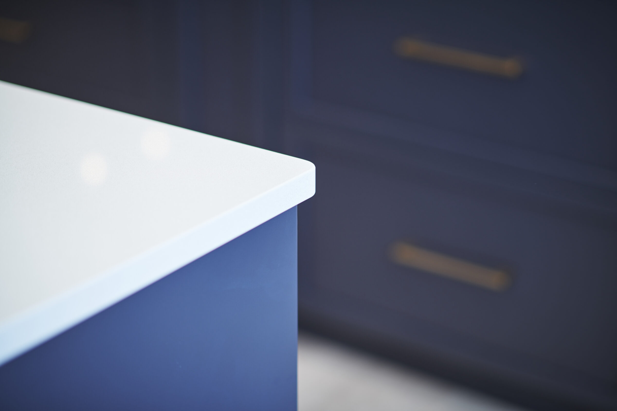 A close-up image focusing on a white countertop corner with a blurred blue cabinet in the background. The cabinet has gold or brass handles.