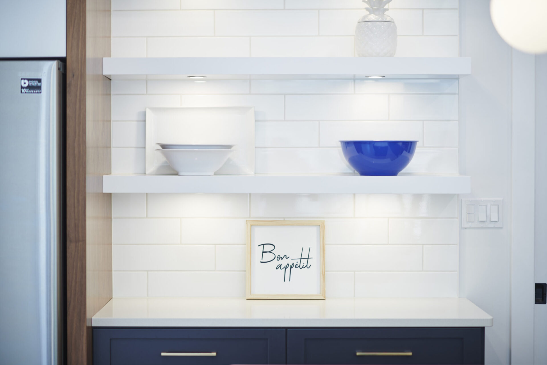 A modern kitchen interior with white subway tiles, two open shelves displaying a blue bowl, a white bowl, and a framed 