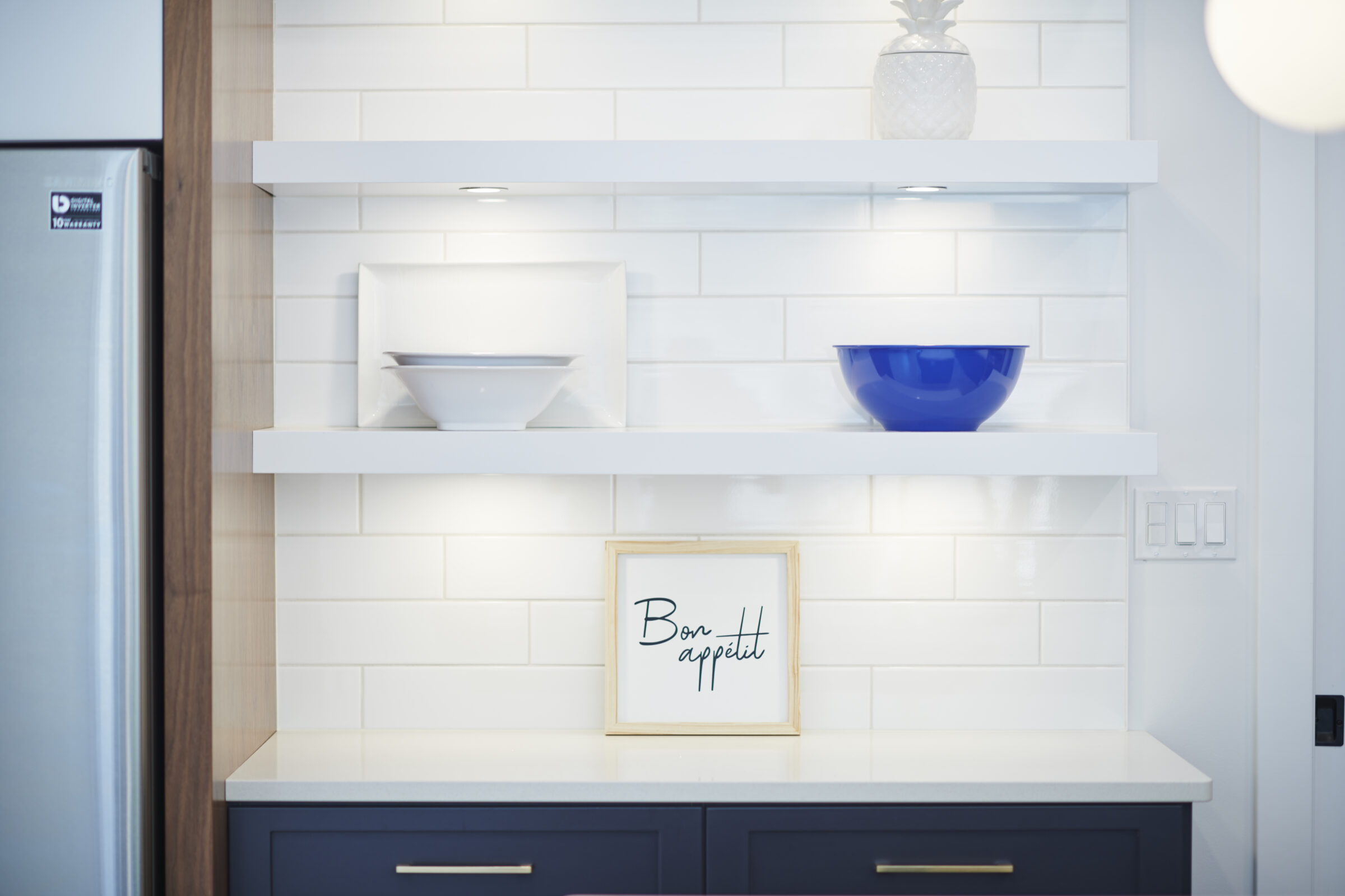 A modern kitchen interior with white subway tiles, two open shelves displaying a blue bowl, a white bowl, and a framed "Bon appétit" sign.