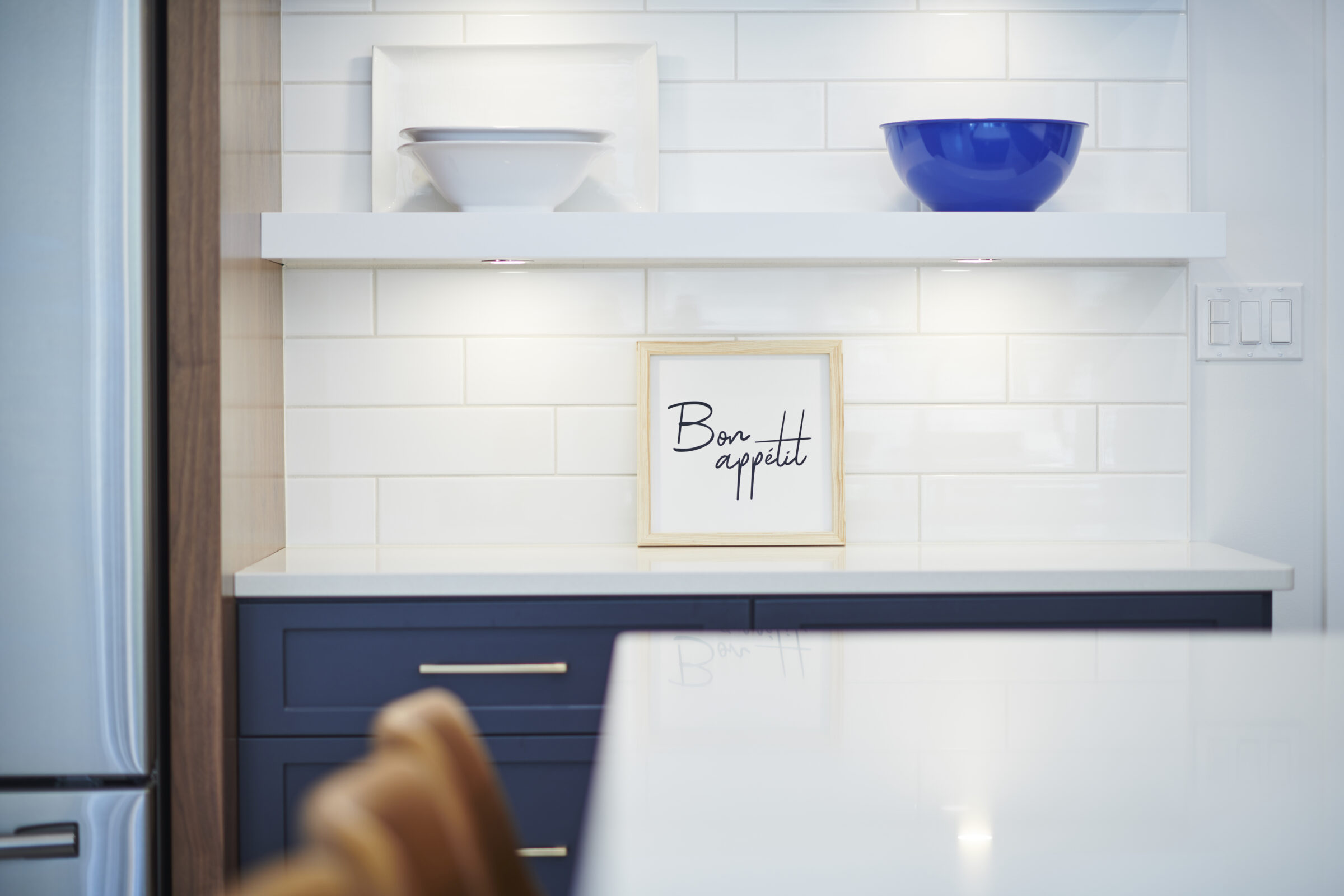 Modern kitchen interior with white subway tiles, floating shelves displaying white and blue bowls, and a framed sign saying "Bon appetit."