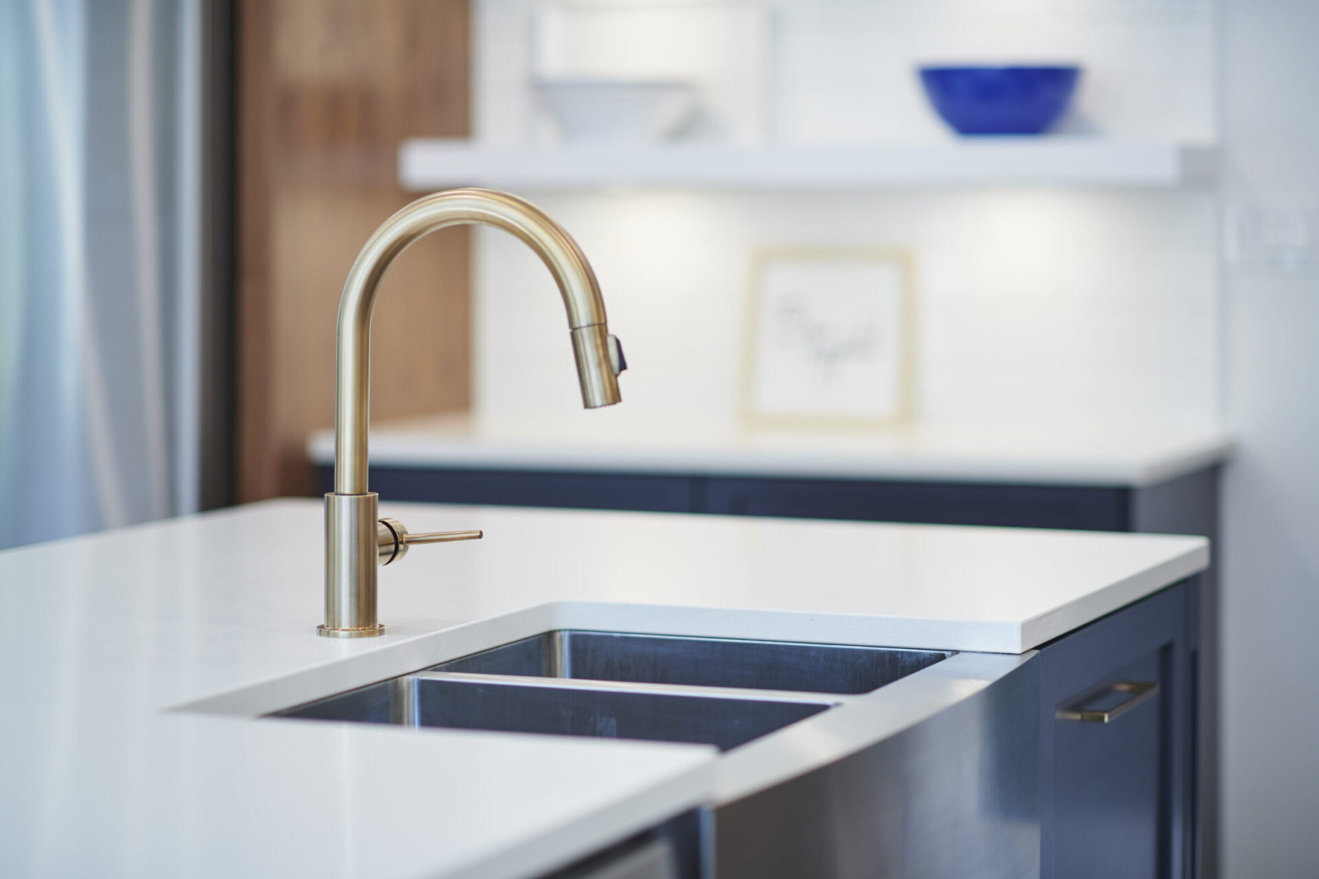 Modern kitchen sink with a brushed gold faucet centered in frame, countertop in focus, with blurred white and navy cabinetry in the background.