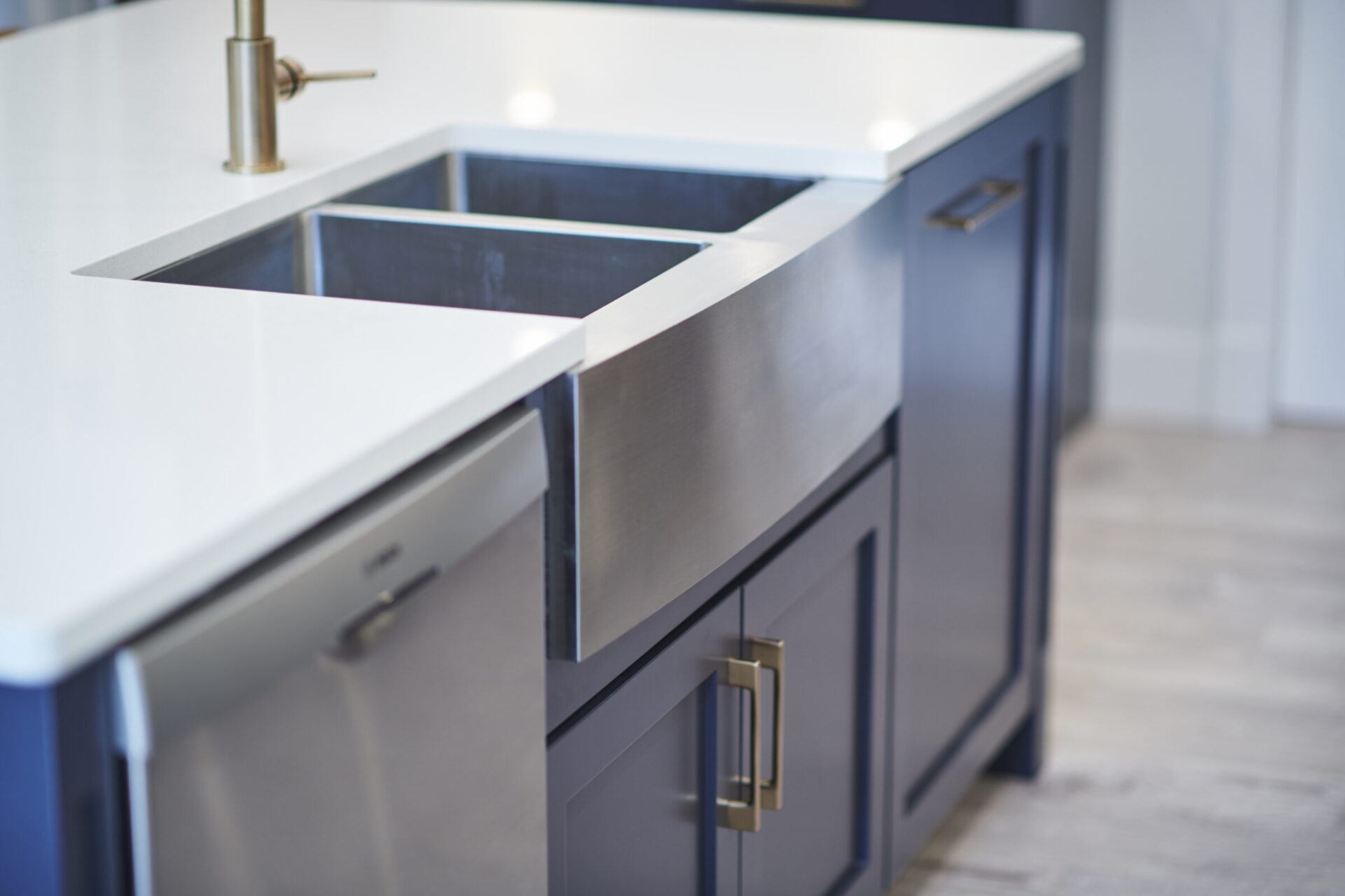 A modern kitchen with a stainless steel undermount sink, white countertop, gold faucet, and navy blue cabinets with brass handles.