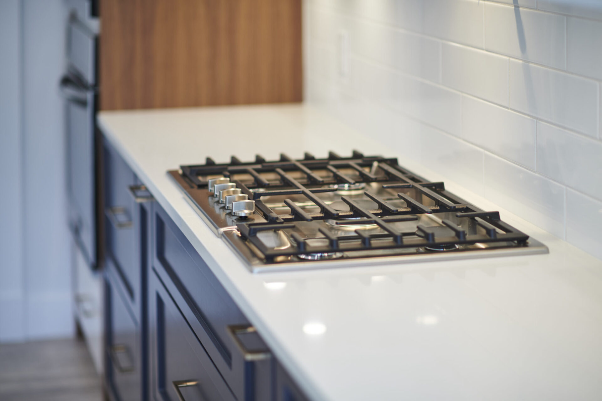 Modern gas stove top with control knobs, set in white countertop. Stainless steel backsplash, wood cabinets, blue drawers, and neutral flooring visible.