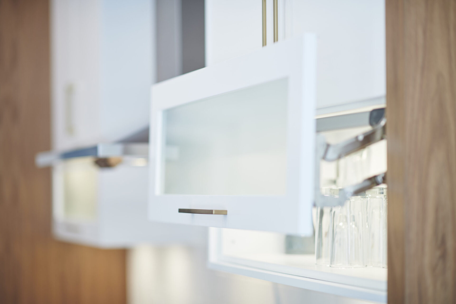 This is a blurred image of an open, modern kitchen cabinet with clear glasses visible, showcasing a minimalist design with wooden accents and white colors.