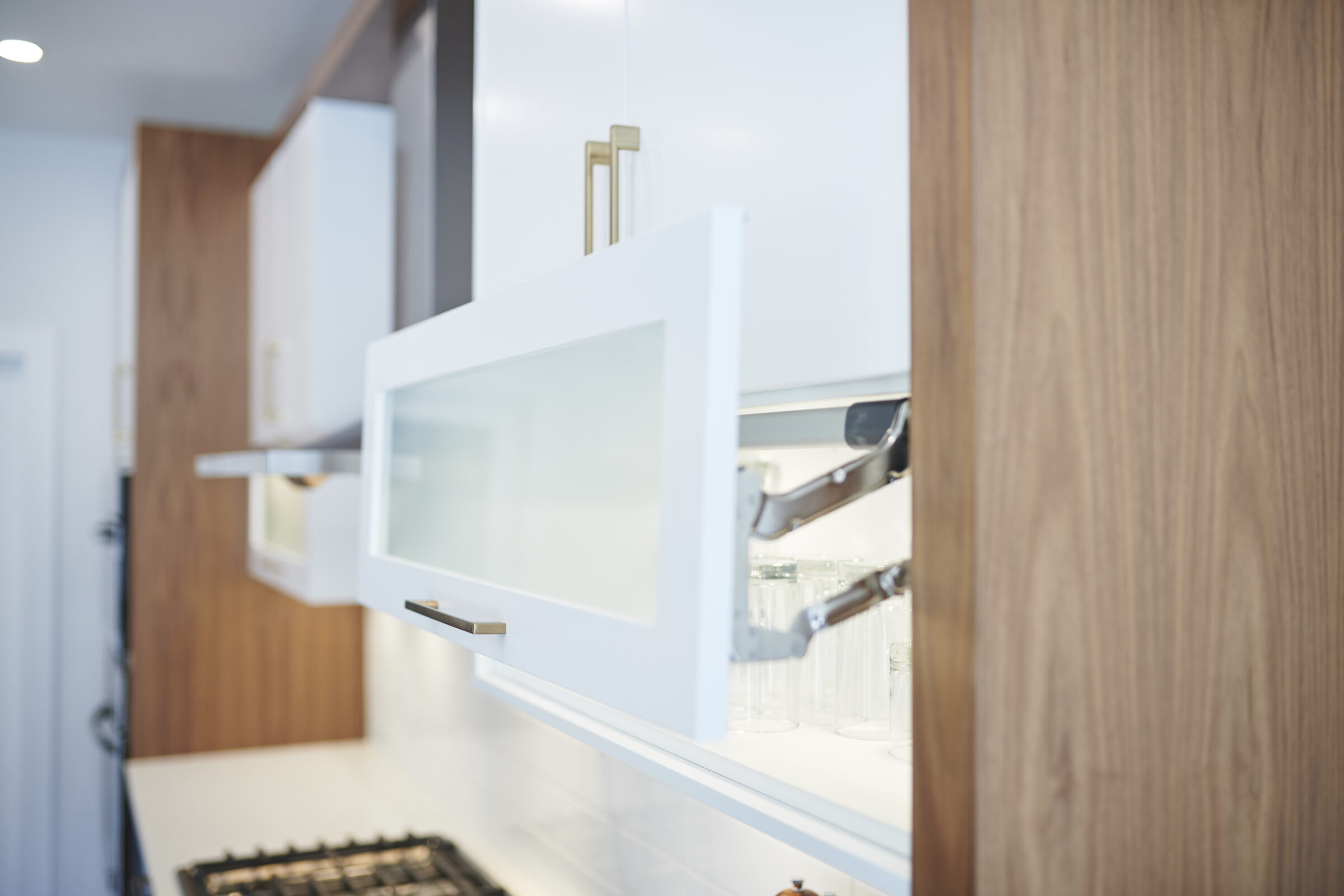 This image shows a modern kitchen with open white cabinets displaying glasses, featuring wooden textures and a blurred gas stove in the background.